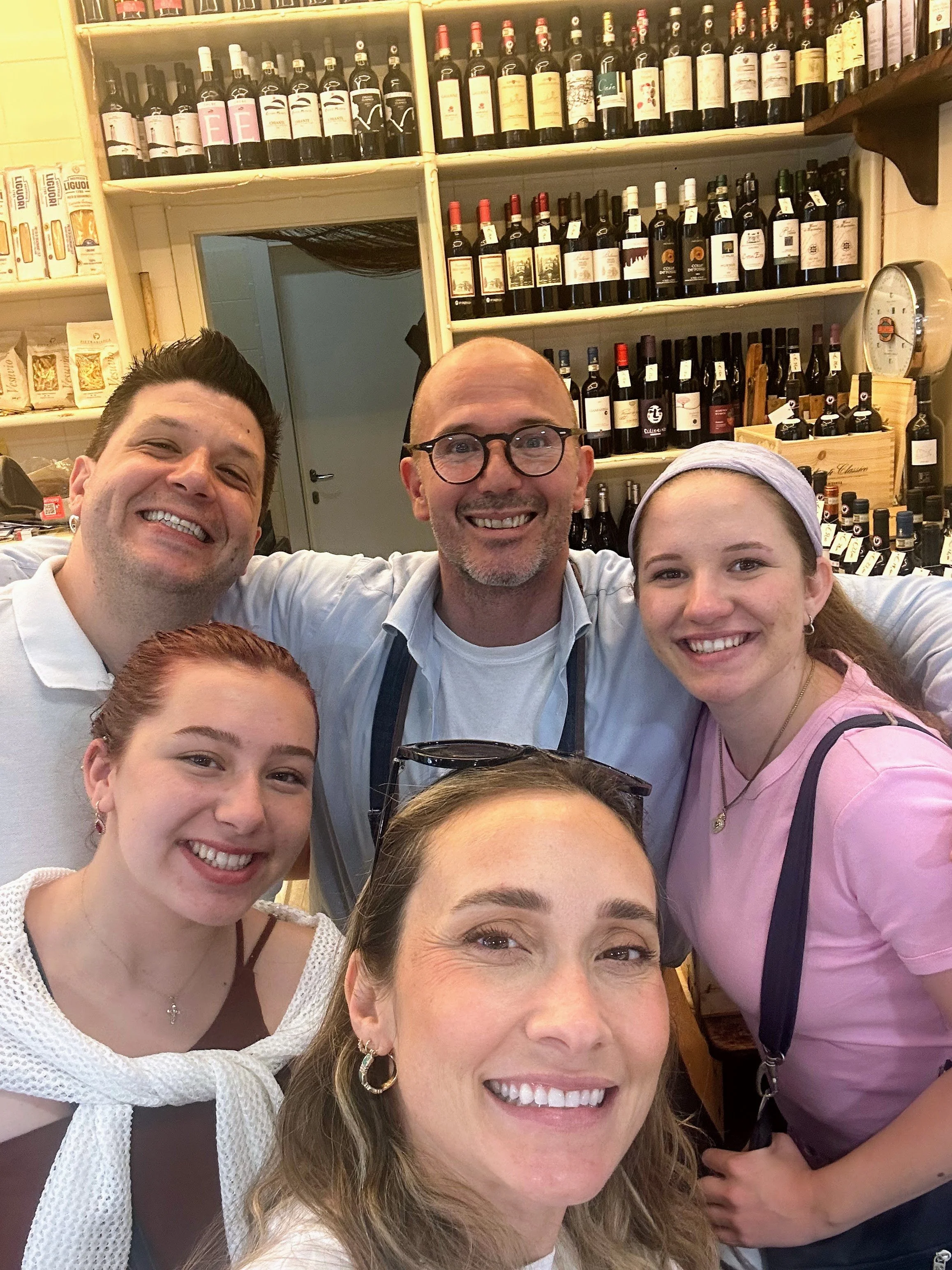 Group of six people smiling for a selfie in a wine shop with shelves of wine bottles in the background.
