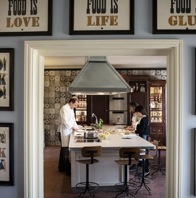 Three people are working in a kitchen, preparing food on a white island counter. They are focused on their tasks, with cabinets and patterned wallpaper in the background. The scene is framed by a doorway with framed wall art above.