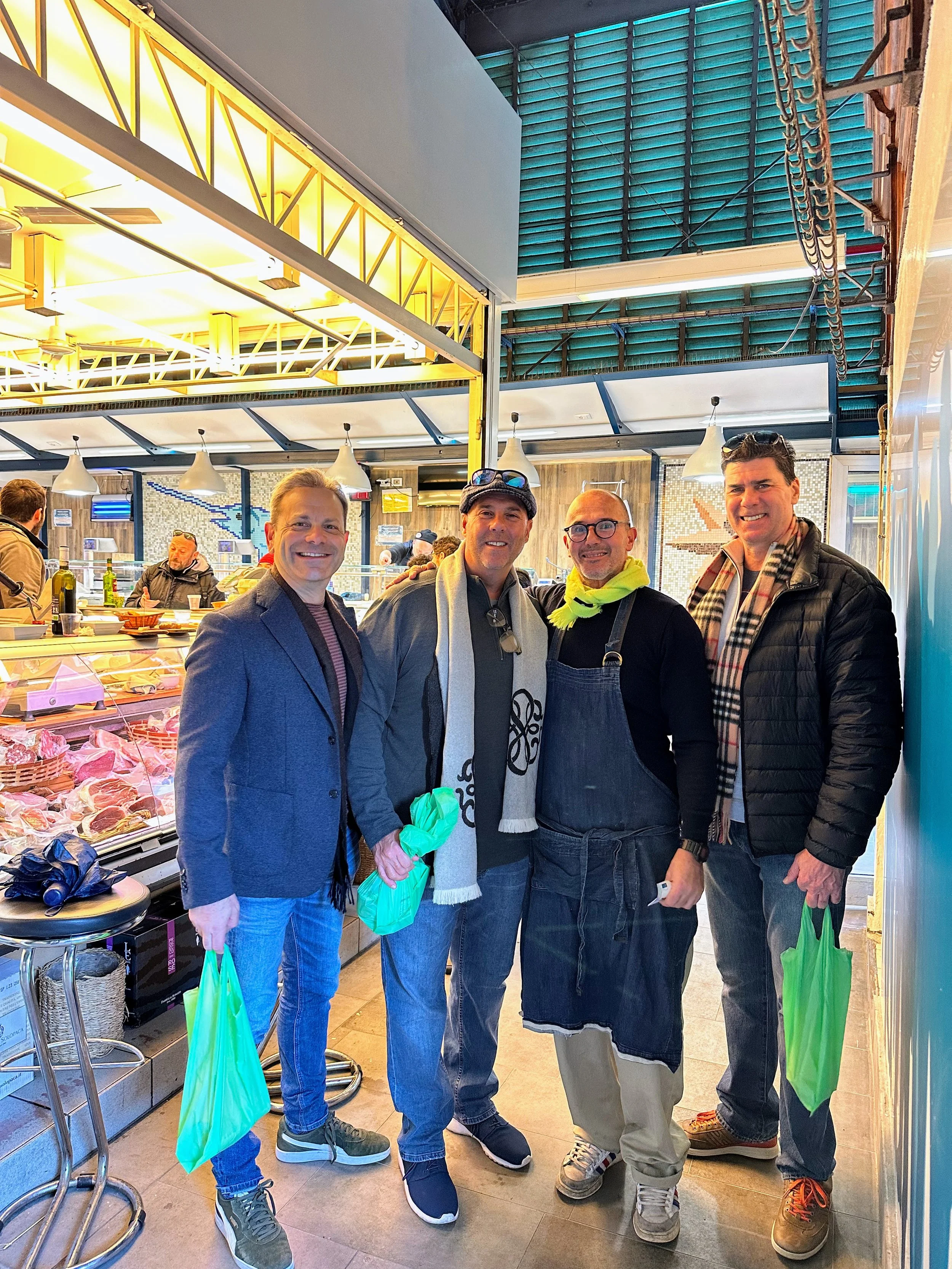 Four men smiling and posing for a photo inside a butcher shop, with meat displayed behind them. They are wearing casual and business casual attire, with some holding green shopping bags. The shop has bright lighting and industrial-style decor.