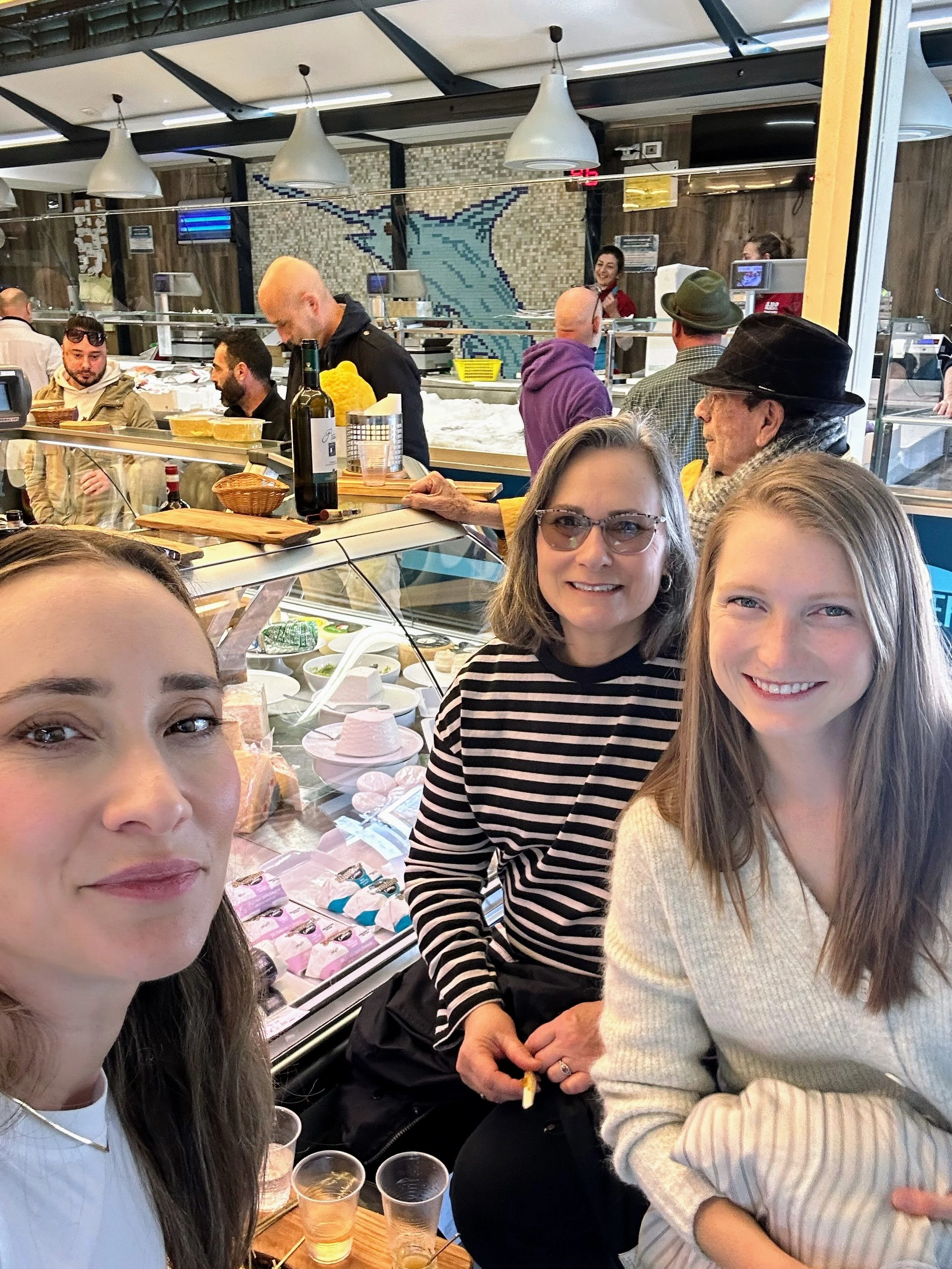 Left front woman with long brown hair, right front woman with long blonde hair, and middle woman with short gray hair and glasses, smiling at a cheese shop counter with view of deli case and people in the background.