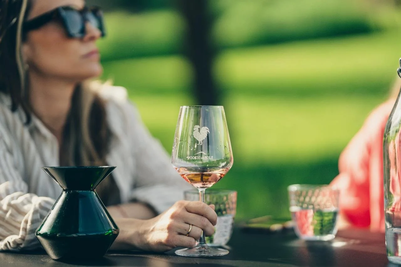A woman wearing sunglasses sitting outdoors at a table, holding a wine glass with pink wine, with a etched rooster and the words "POCHETTE A BOHEME" on the glass. There are also two glasses of water and a black vase on the table. The background is bl