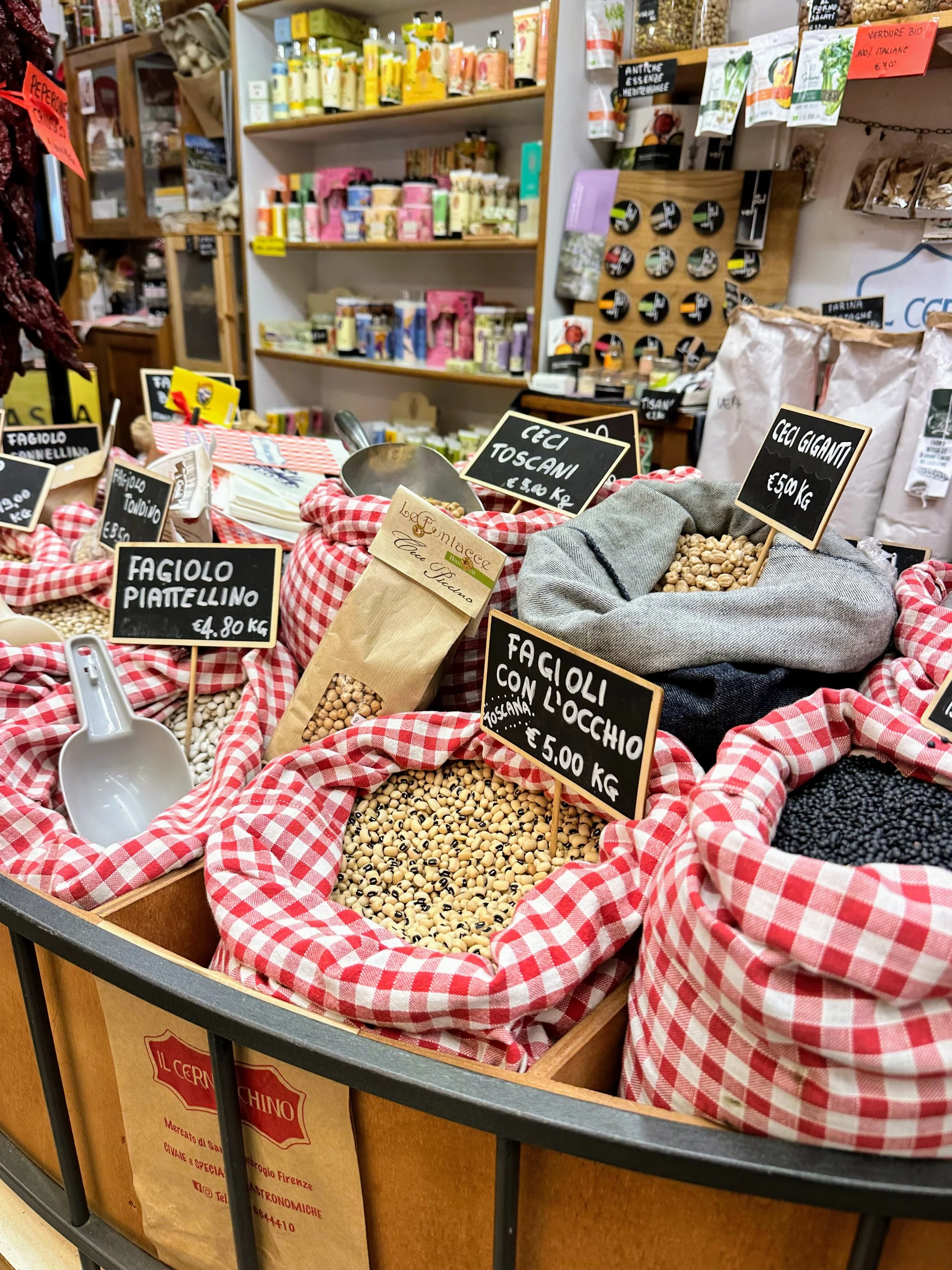 Assorted dried beans and legumes in cloth-lined bins with handwritten signs displaying prices, including packages labeled 'Fagioli Piattellino,' 'Ceci Toscani,' and 'Fagioli con l'occhio,' on display in a grocery store