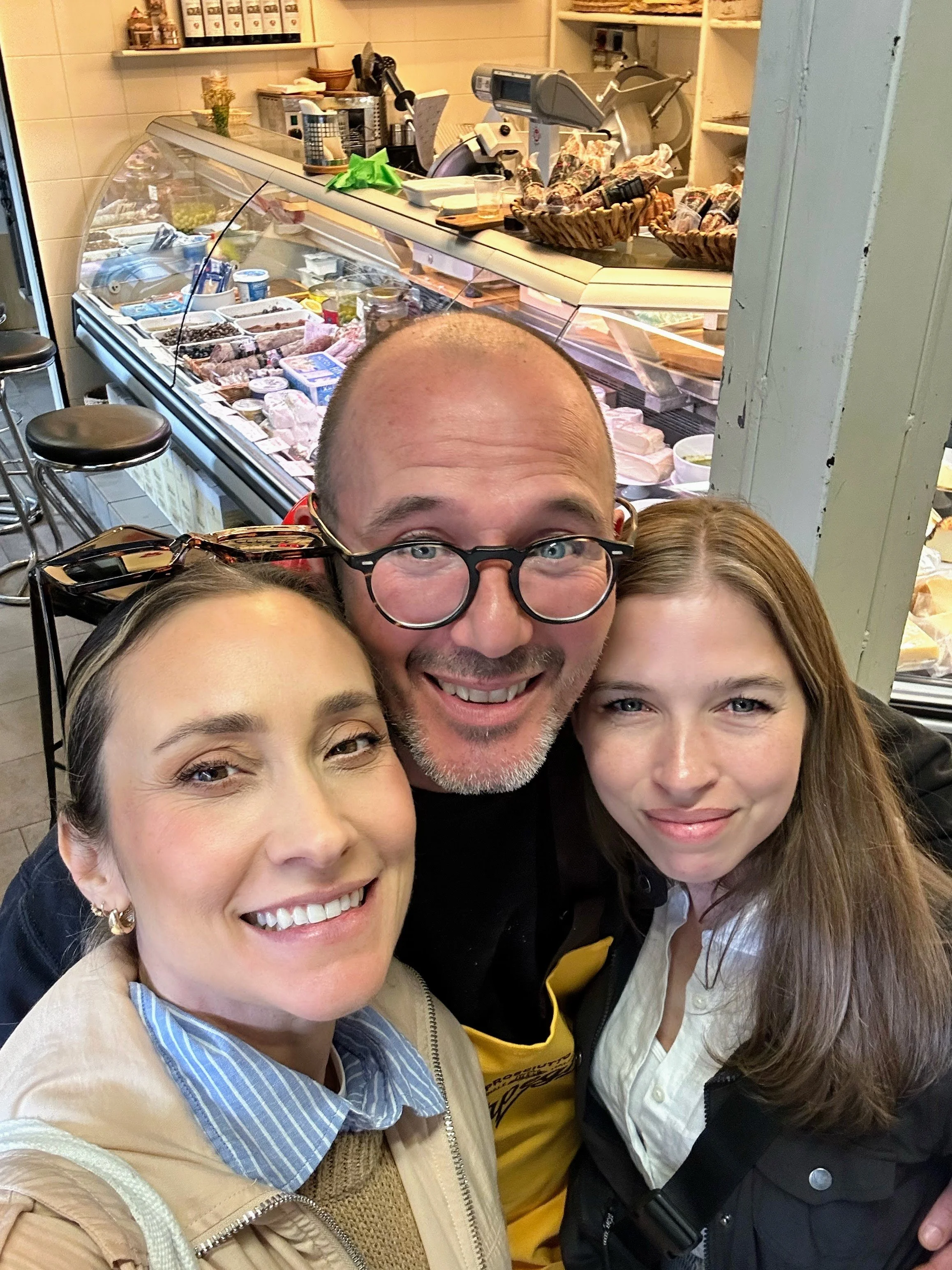 Three smiling people taking a selfie in front of a display case filled with various cheeses, meats, and deli items inside a shop or café.