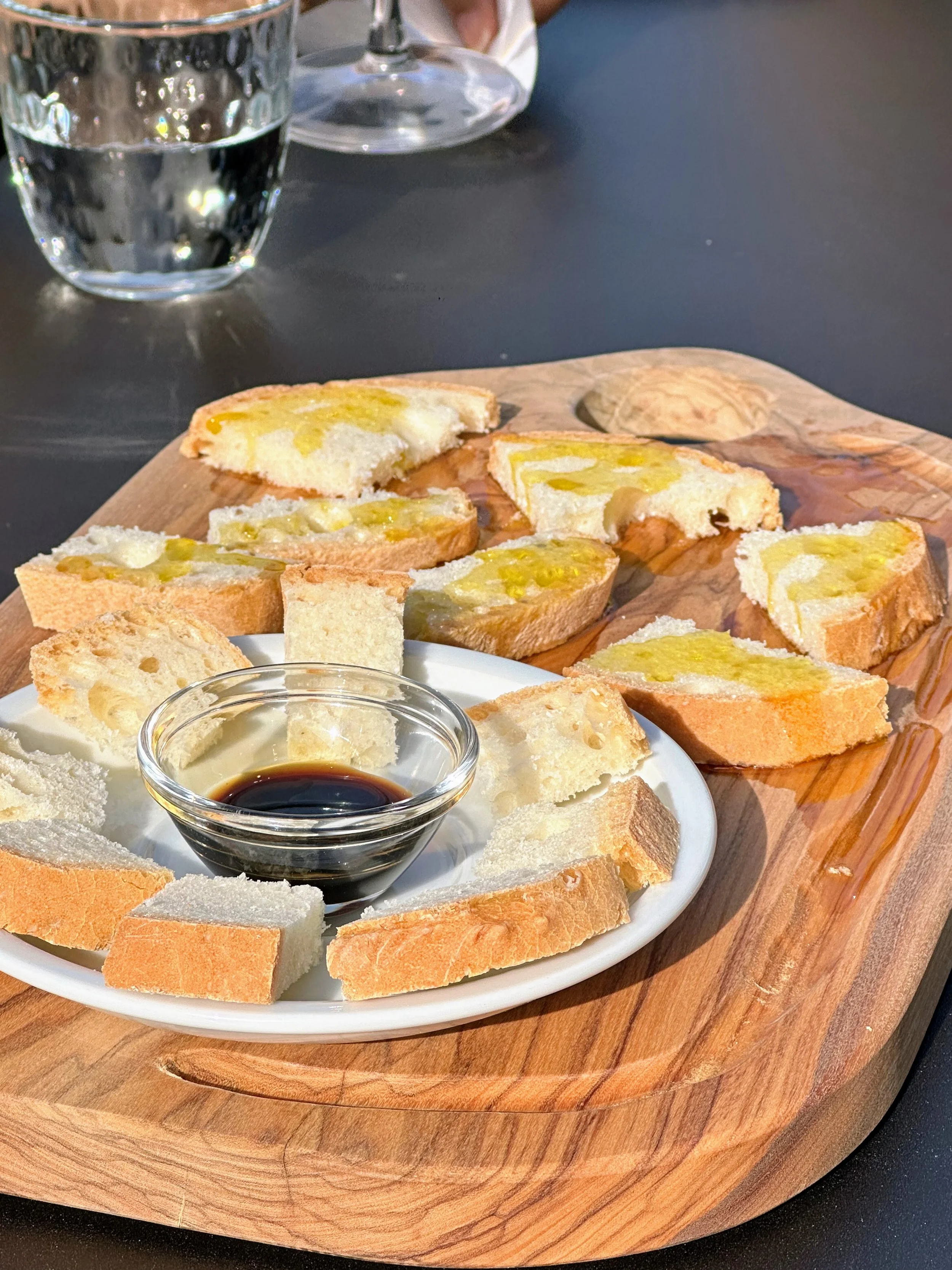 Slices of bread with olive oil and balsamic vinegar on a wooden serving board, with a glass of water in the background.