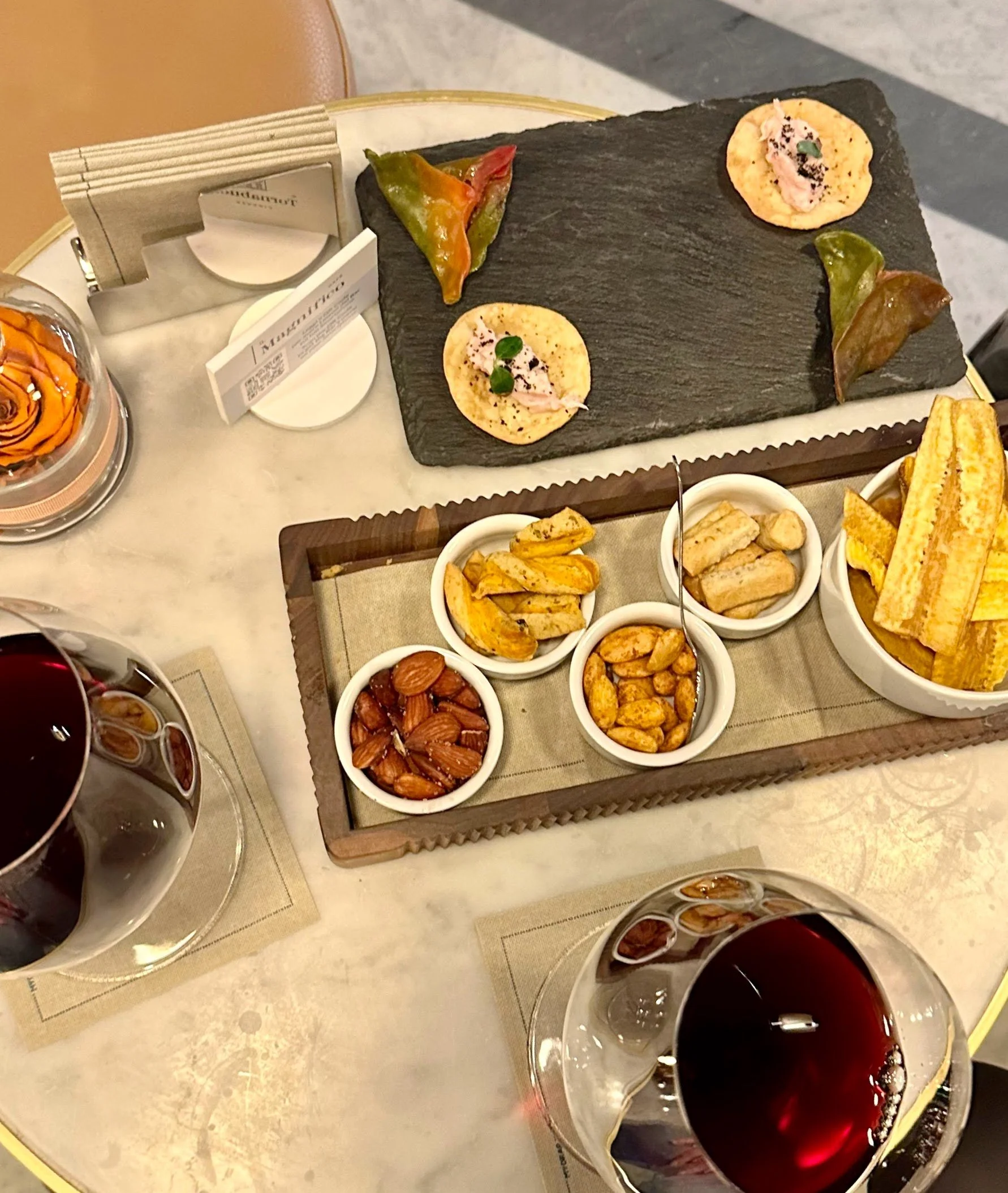 A table with a variety of appetizers, including nuts, plantains, and cheese chips, along with two glasses of red wine, and a slate serving board with small decorated snacks. There are also utensils and small menus or notes on the table.