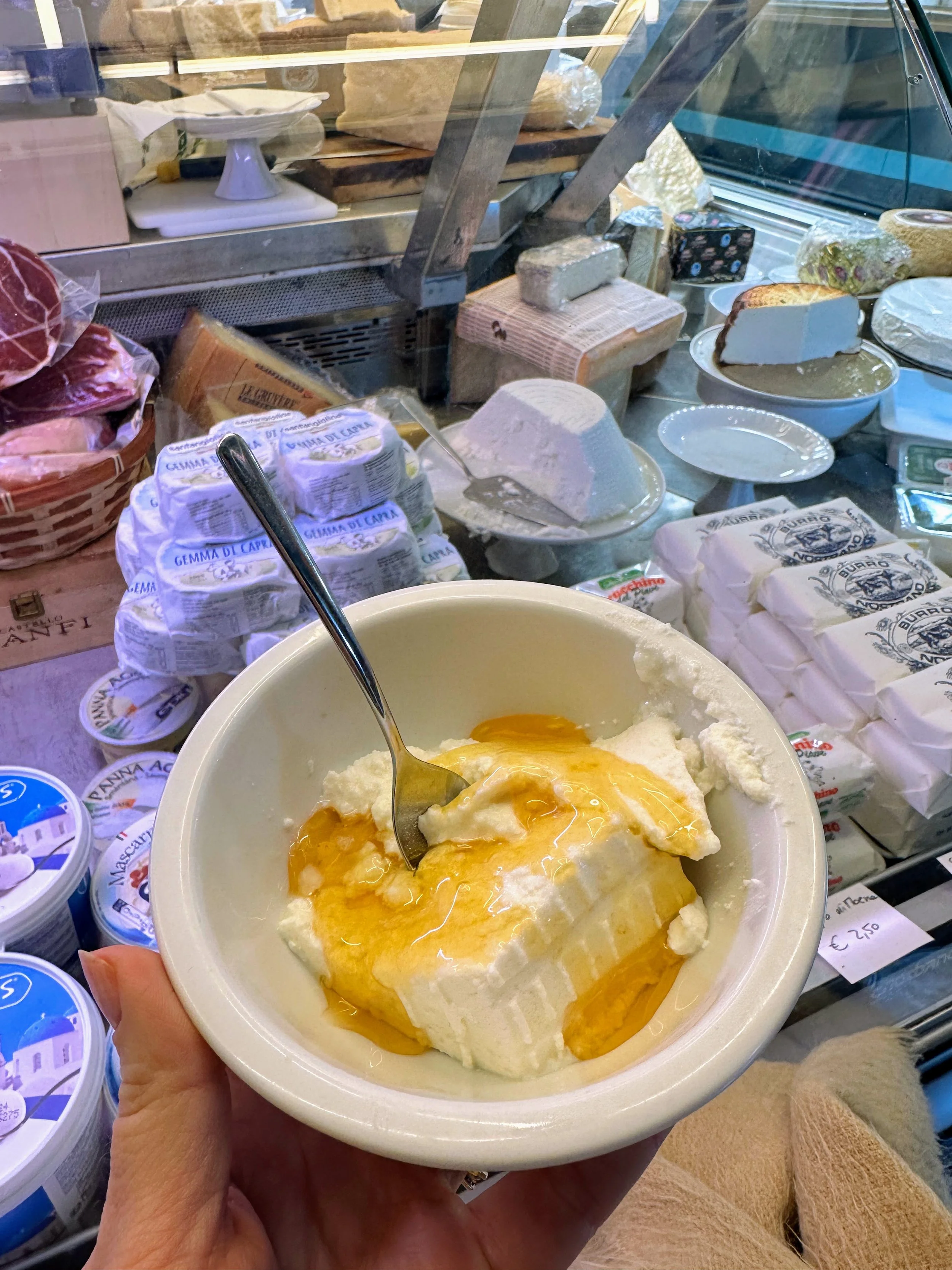 Hand holding a bowl of ricotta cheese topped with honey, in front of a cheese display case with various cheeses and dairy products.