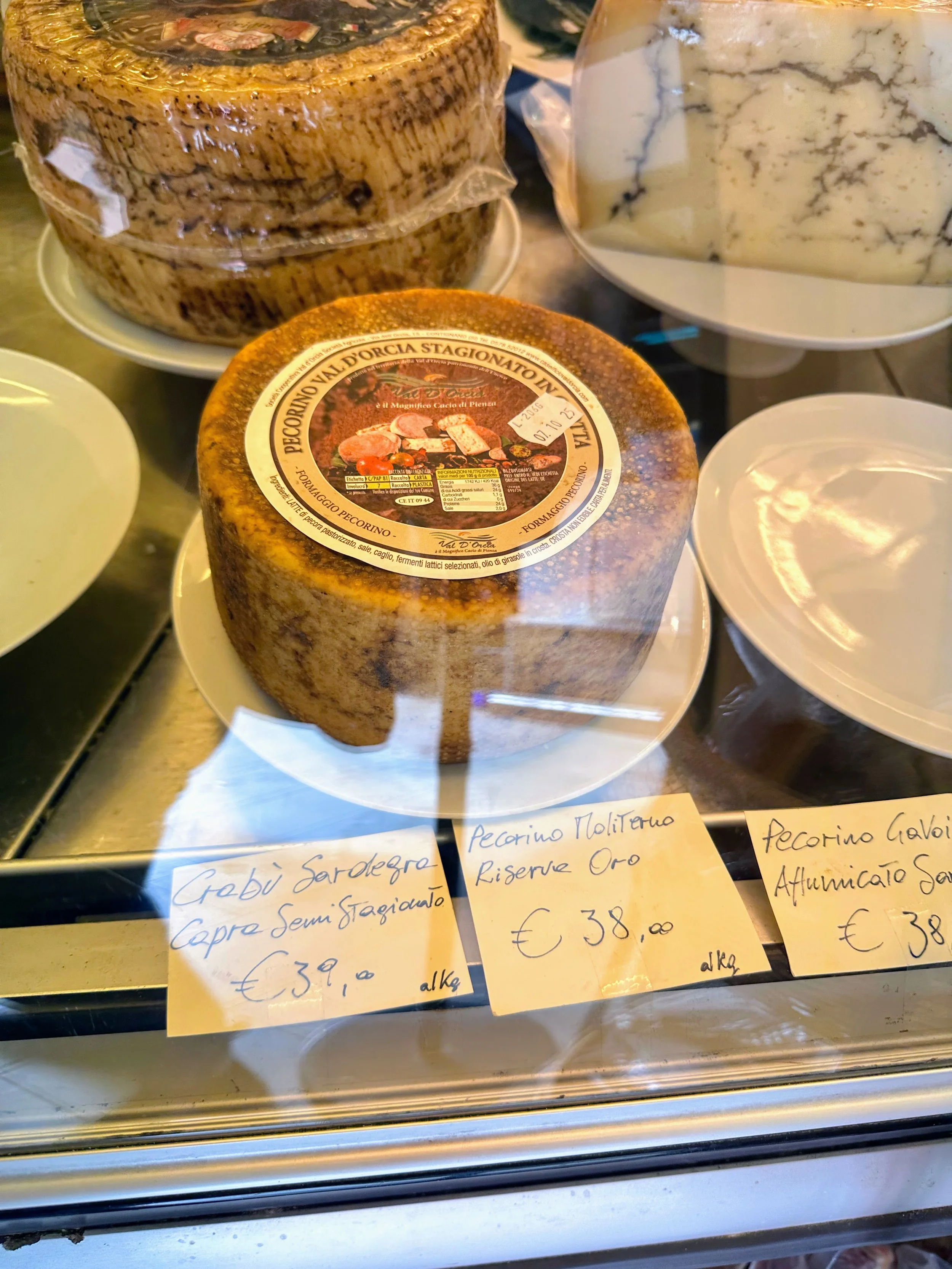 Display of various cheeses, including Pecorino, with handwritten price tags in Italian. The cheeses are behind a glass display.
