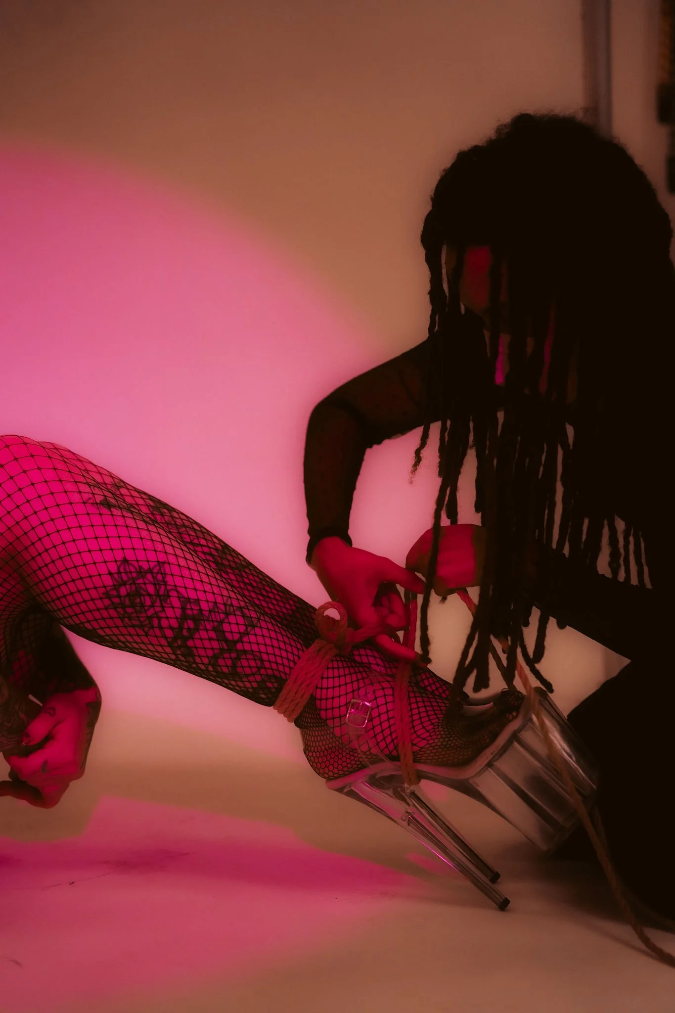 A person with long locks wearing black fishnet stockings with floral patterns and high heels, posing against a pink-lit background.