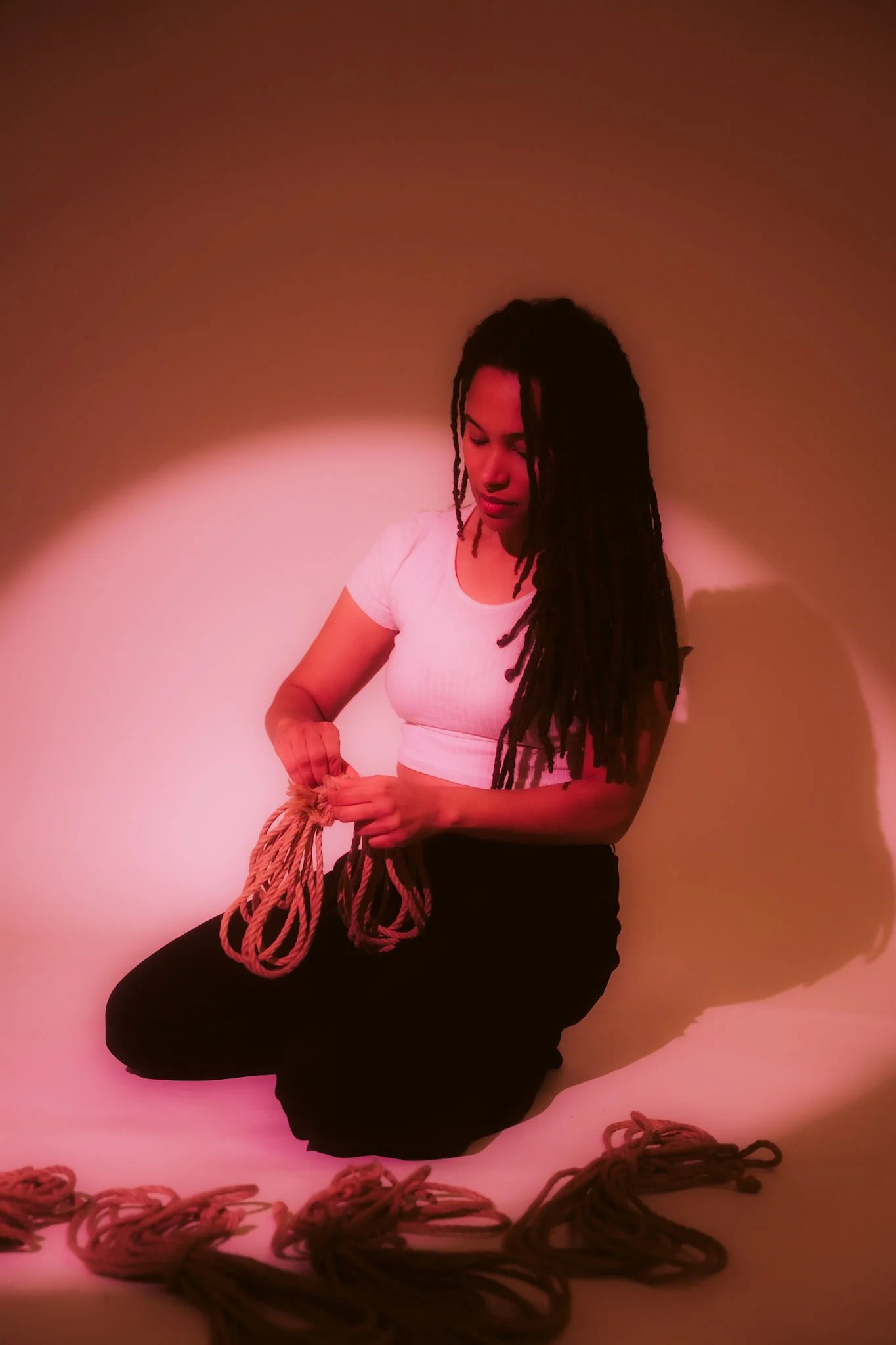 A person with long ocks, wearing a white crop top and black trousers, kneels on the ground in a pink-lit room, holding a piece of rope.