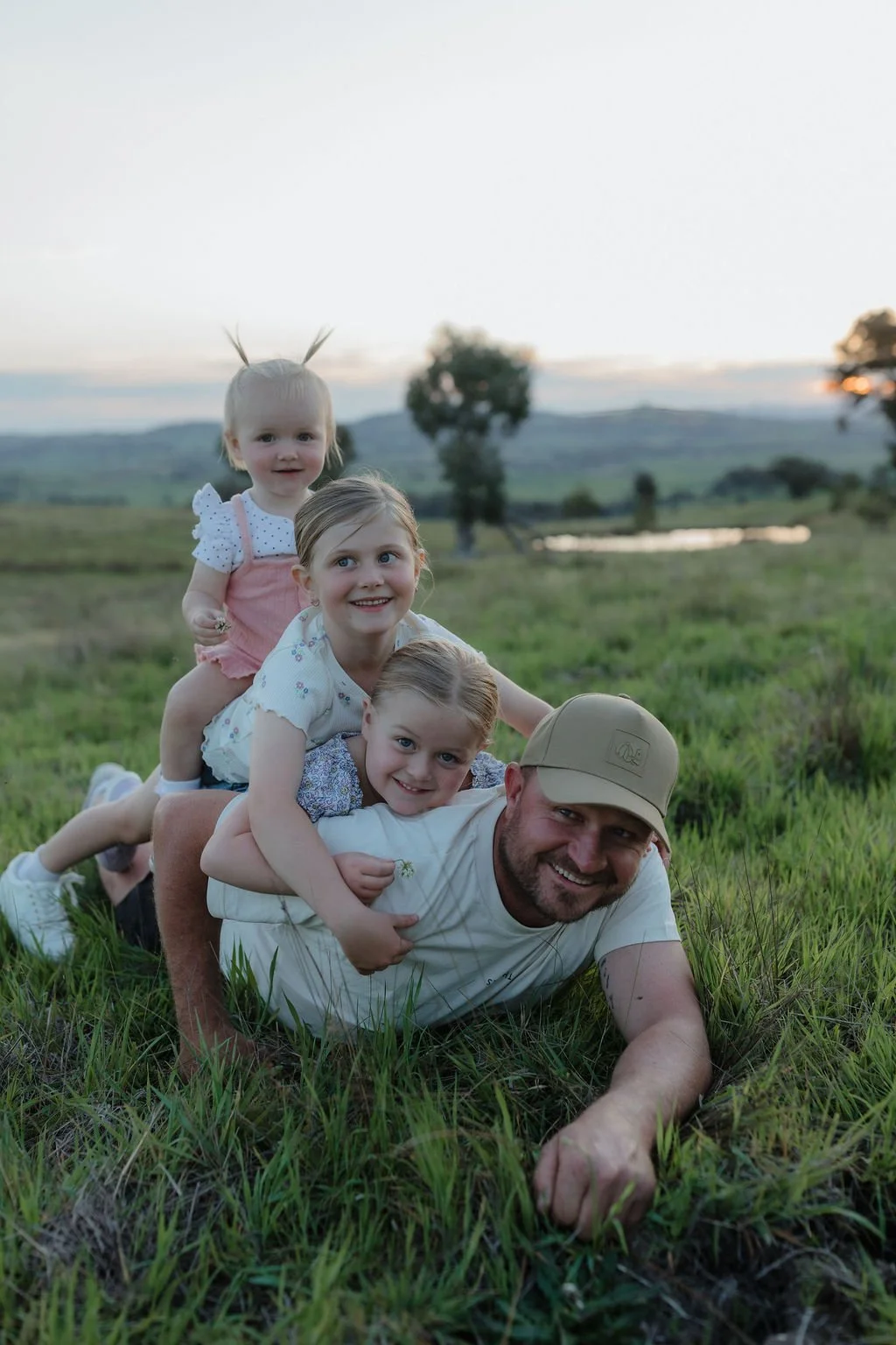Dad lying in the grass with his three daughters piled on top of him during a playful family photoshoot, captured by Jade Jones Photography.