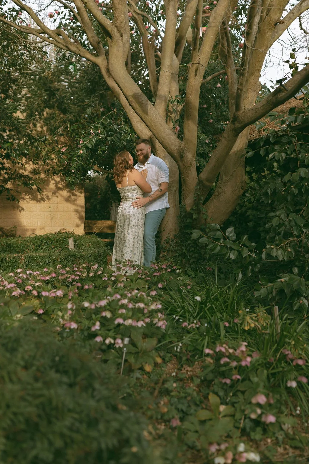 Engaged couple cuddling in a beautiful garden as the man leans against a tree and they look into each other’s eyes during a romantic engagement photoshoot.