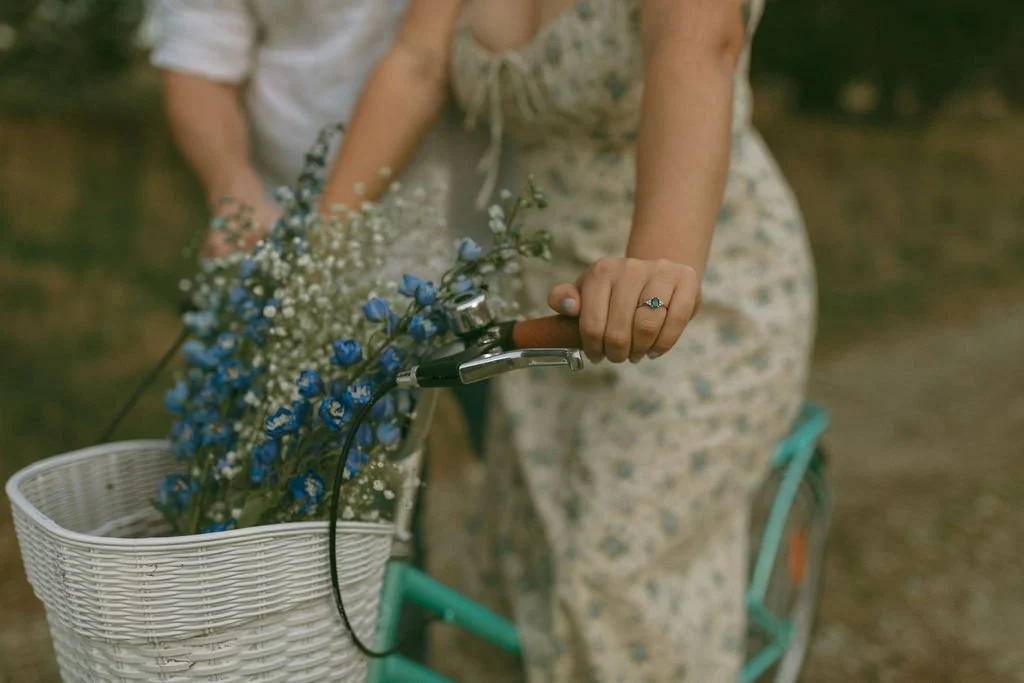 Close-up of an engagement ring as the bride-to-be sits on a vintage bike with flowers in the basket, her fiancé standing beside her during an engagement photoshoot.