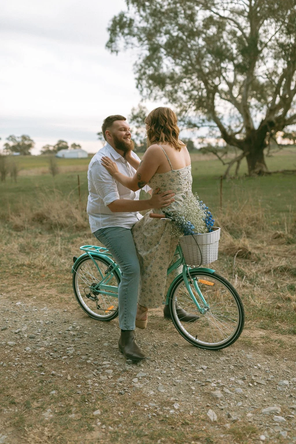bride-to-be sitting on the vintage bike handlebars as her fiancé holds her waist, flowers in the basket and a dreamy farm backdrop.