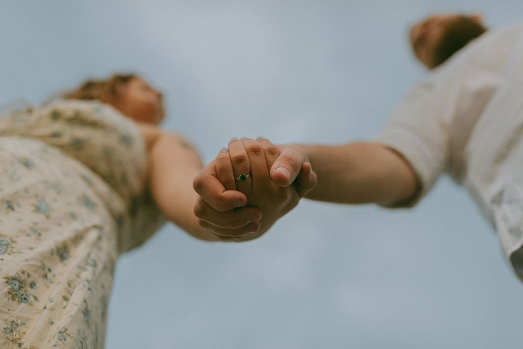 Close-up of an engaged couple holding hands, showcasing her engagement ring with a blurred background, captured by Jade Jones Photography.