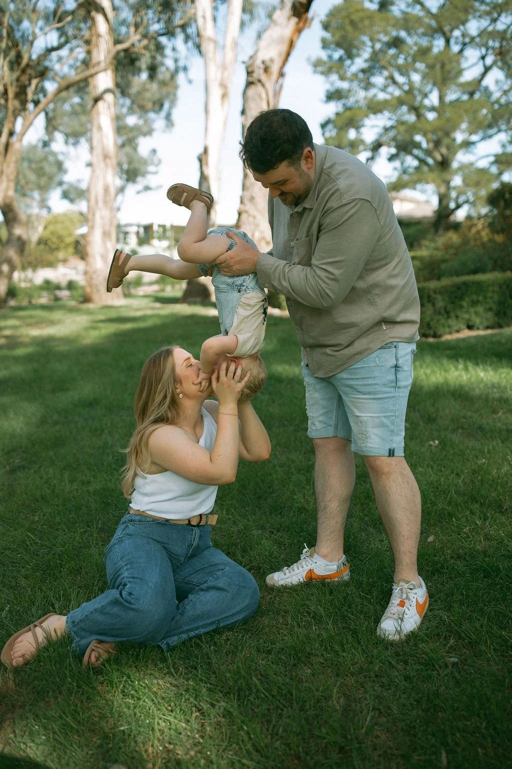 Parents sharing a joyful moment in the park as the dad holds their toddler upside down and the mum kisses him during a relaxed family photography session by Jade Jones Photography.