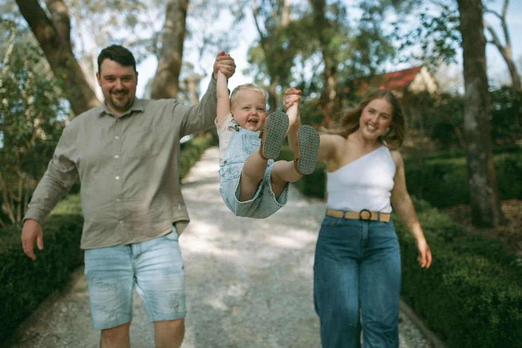 Mum and dad holding hands while swinging their toddler into the air during a relaxed family photography session in the park, captured by Jade Jones Photography.