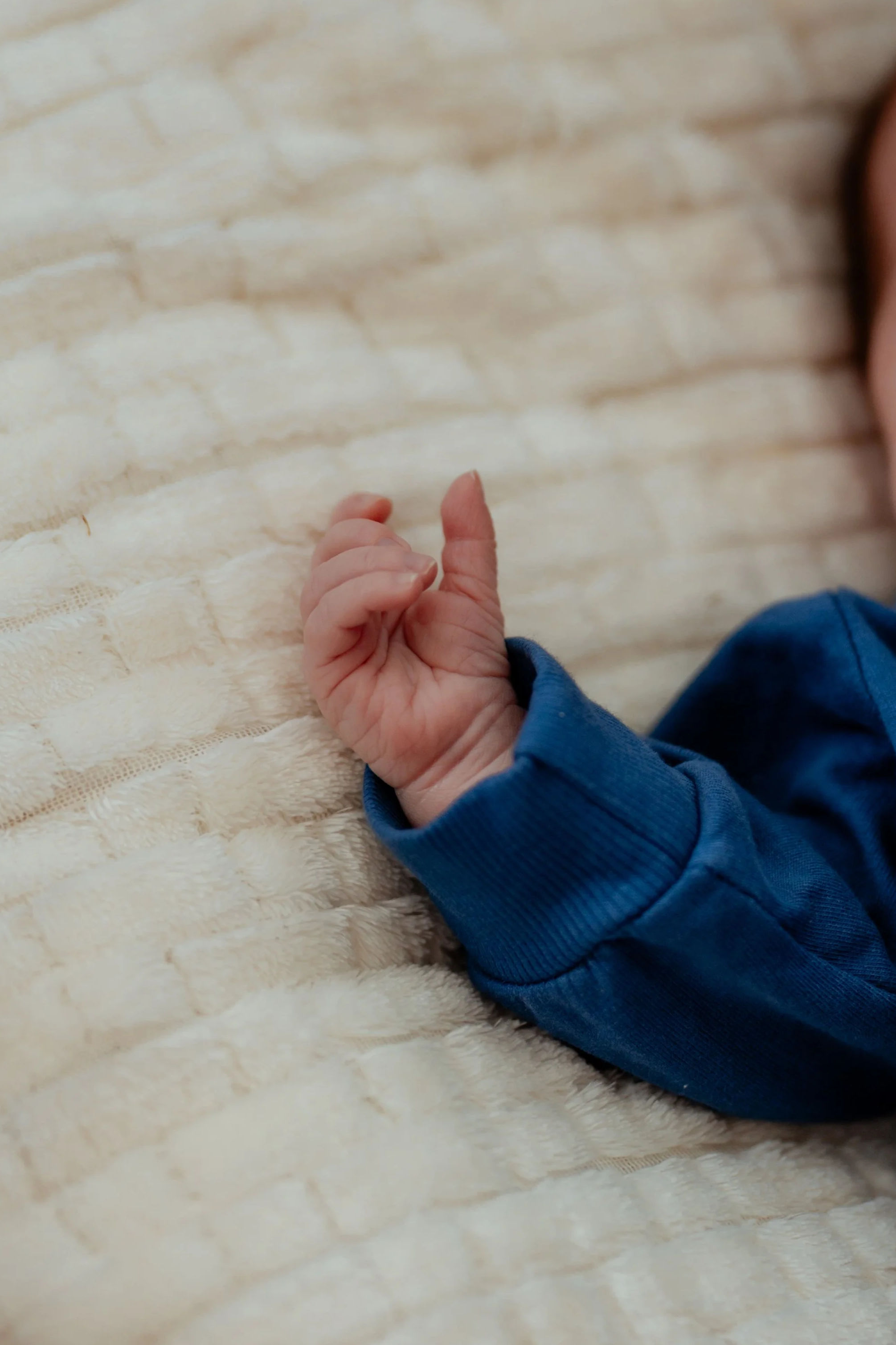 Main d'un bébé reposant sur un lit en tissu beige, vêtu d'une chemise bleue.