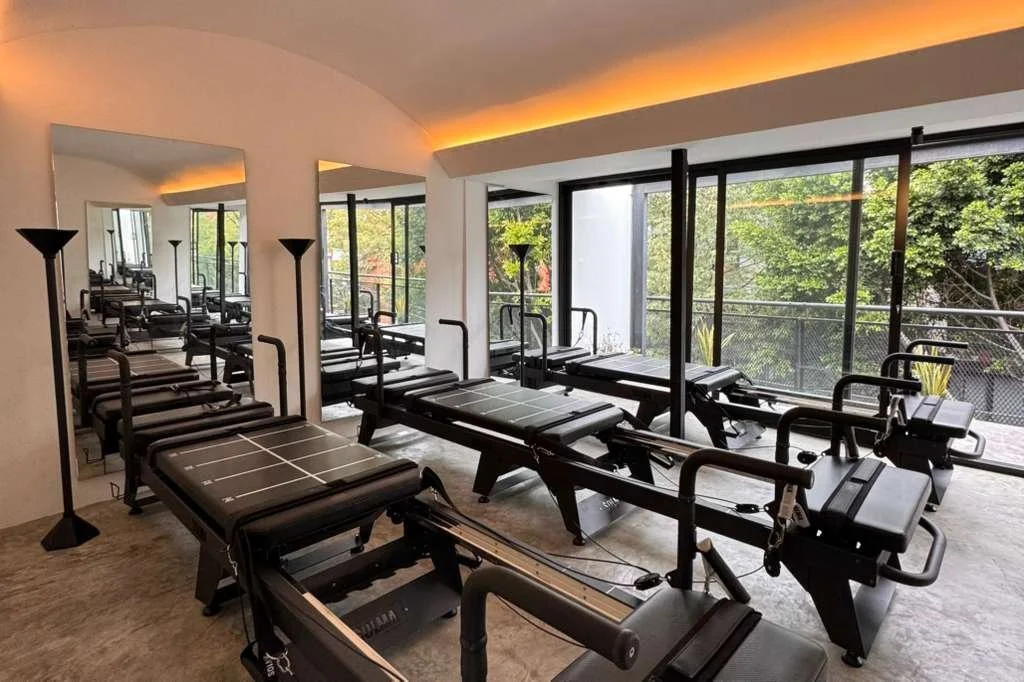 Sala de pilates con varias máquinas de ejercicio frente a ventanas de vidrio con vista a árboles verdes.