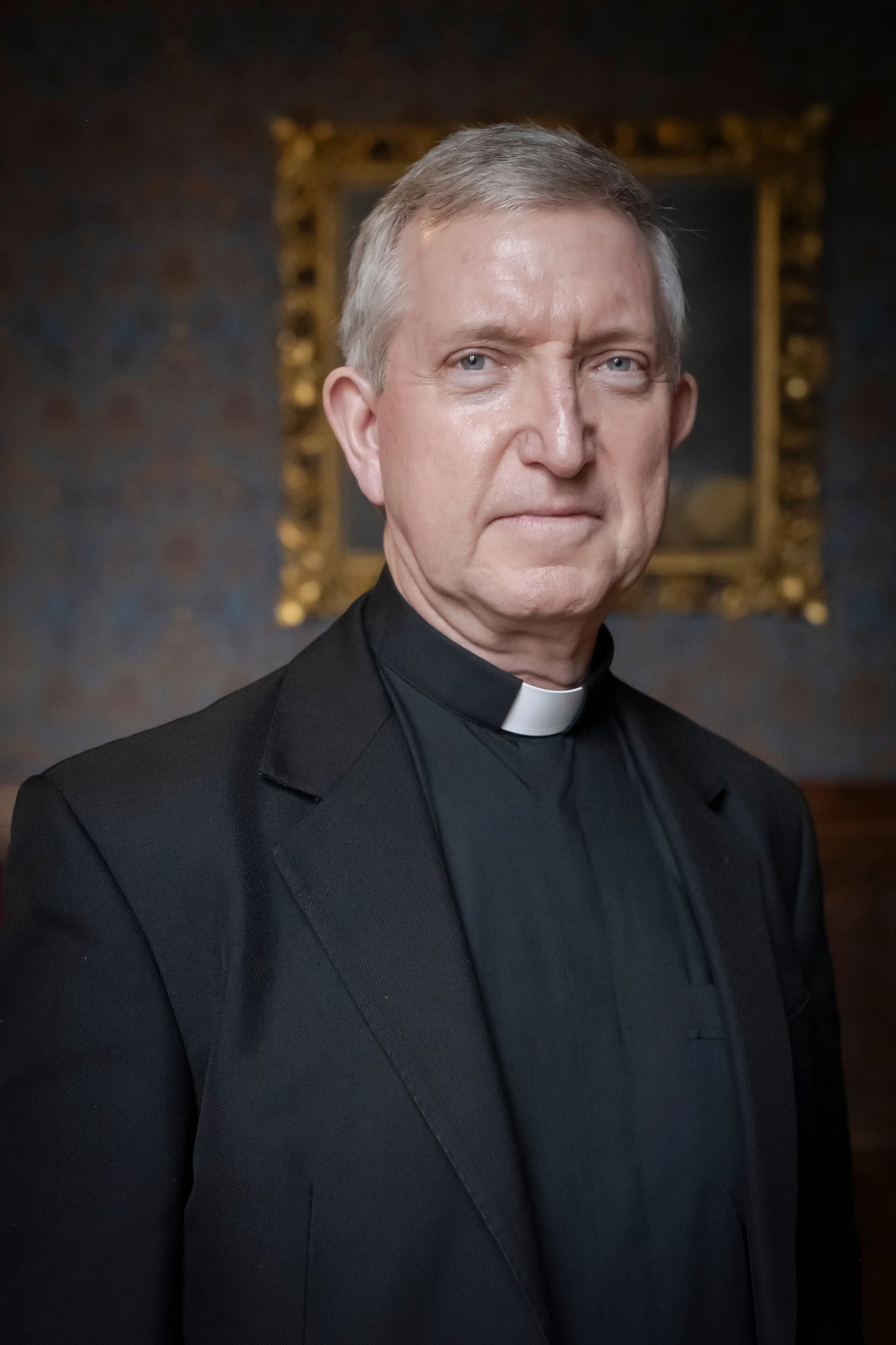 An image of Paul McPartlan dressed as a priest in a black suit with a clerical collar, standing in front of a dark background with a gold-framed painting.