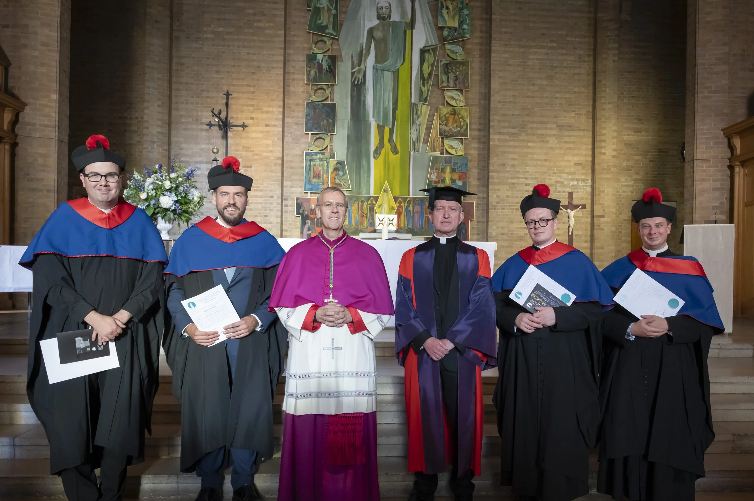 A group of six people in academic regalia standing in front of a religious altar inside a church. The group includes men and women, with some holding certificates. Behind them is a large mosaicked artwork of Jesus Christ, flanked by various smaller depictions of religious scenes, and a floral arrangement is placed to the left.