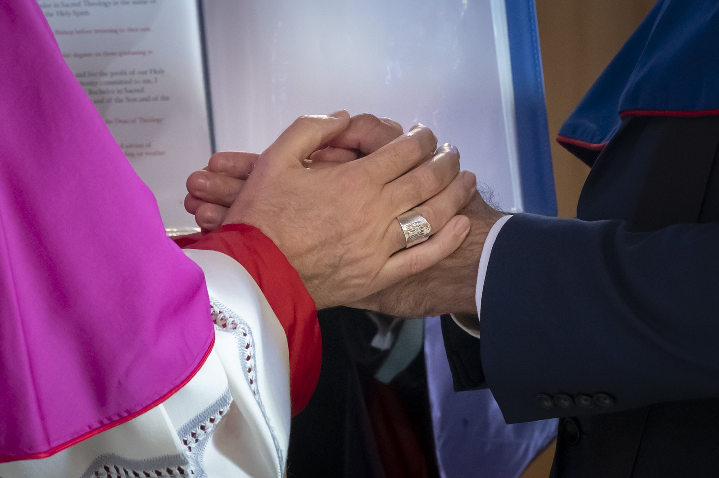 A person in a dark suit with a white shirt is holding the hand of a woman wearing a pink dress with a white and red embroidered shawl. The background shows a presentation or display board with text.
