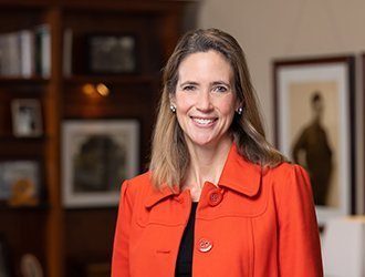 A smiling woman with light brown hair in a red blazer in an indoor setting with pictures on the wall behind her.