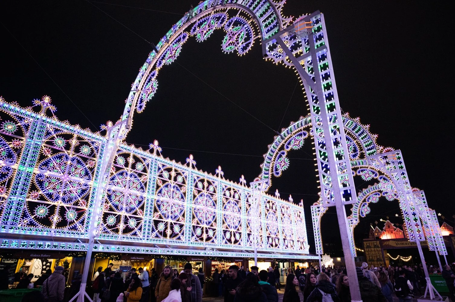 Nighttime scene at a festive holiday fair with illuminated decorative arches and structures, crowds of people walking and shopping.