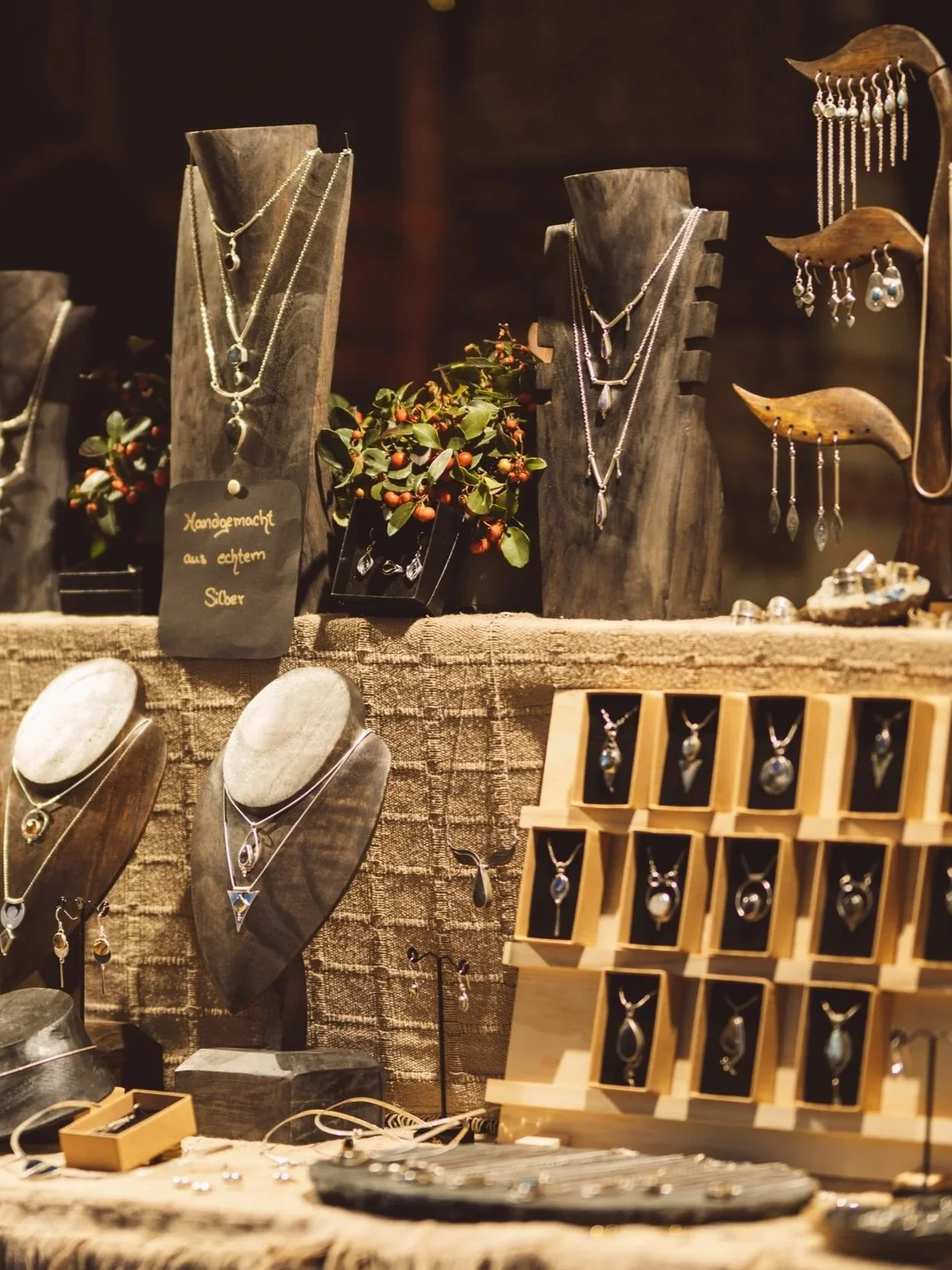 Jewelry display table with necklaces and earrings on wooden stands and trays, decorated with small plants and a handwritten sign in German