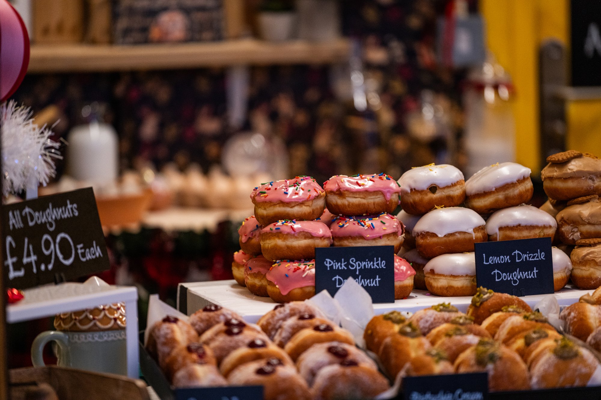 Display of various donuts with pink, white, and brown icing, topped with sprinkles and other toppings, arranged in rows at a bakery or donut shop.