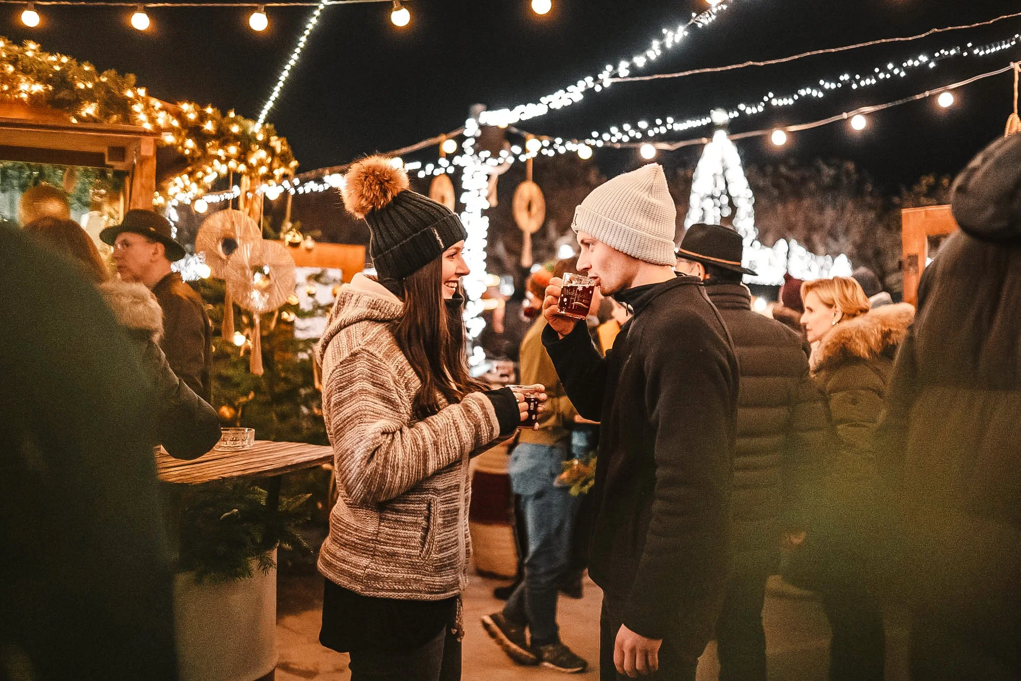 A young woman and young man standing face to face at a festive outdoor gathering during winter, sharing a smile, with the woman holding a drink and the man sipping from a cup, under string lights and holiday decorations.