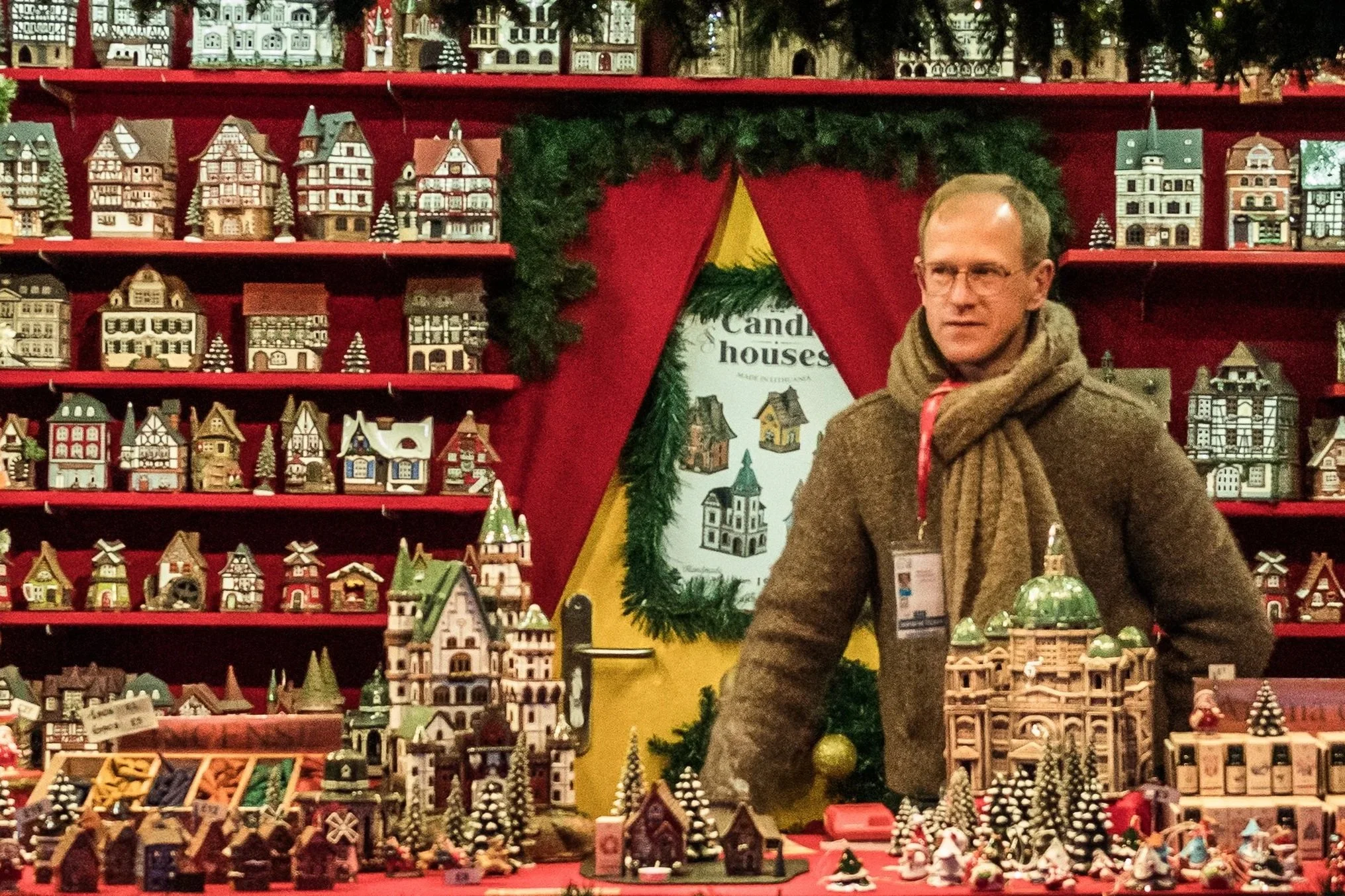 Display of various miniature wooden houses and Christmas decorations on red shelves, with a man standing in front of the display.