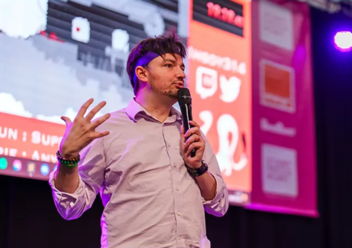 Un homme en chemise blanche parle en tenant un micro lors d'une conférence ou d'un événement, avec un grand écran en arrière-plan affichant du contenu numérique.