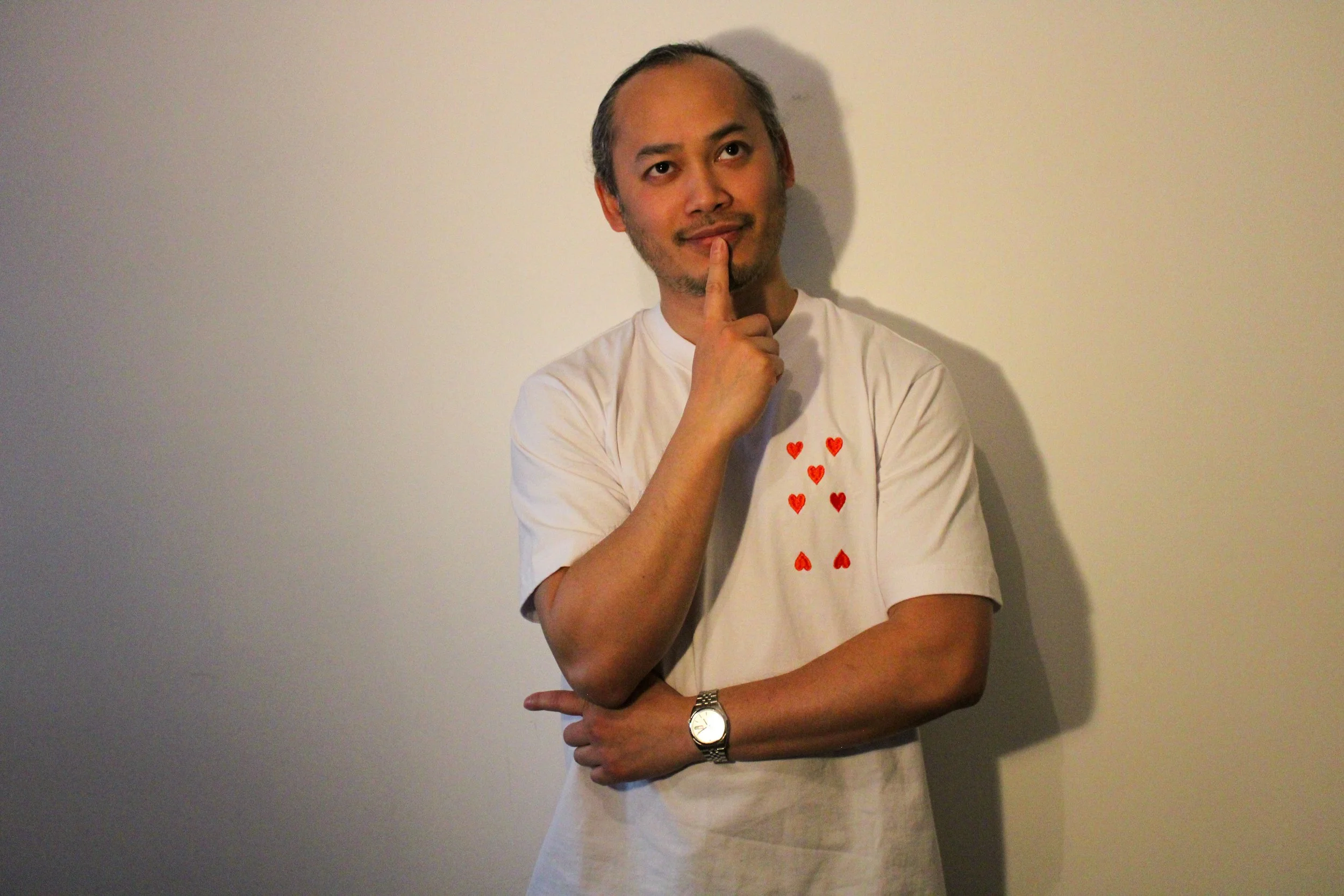 Homme pensif portant un t-shirt blanc avec des coeurs rouges, debout contre un mur beige, faisant un geste de réflexion avec le doigt sur le menton