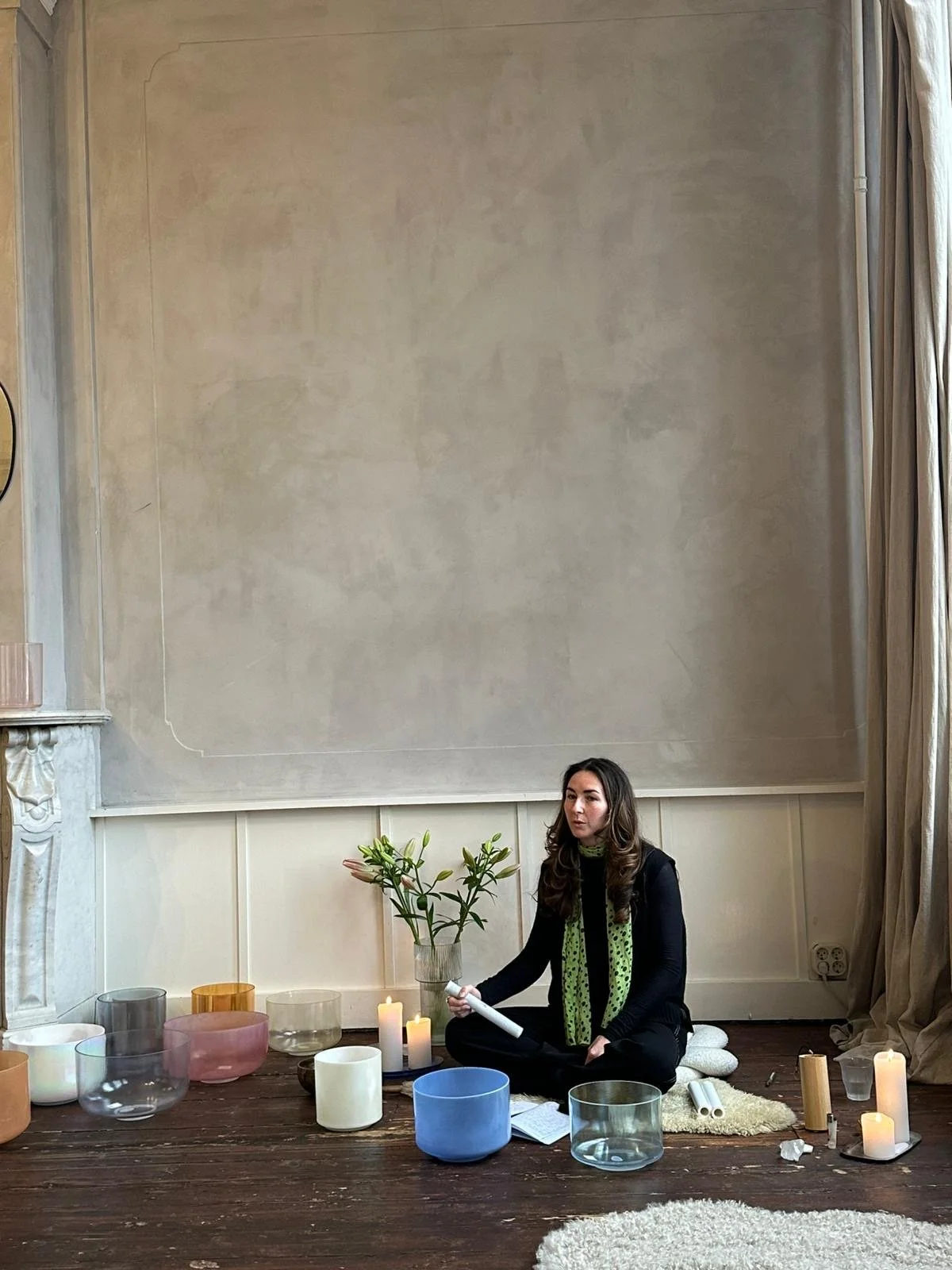 A woman sitting on a wooden floor surrounded by singing bowls, candles, and plants in a decorated room facilitating breathwork for a team.