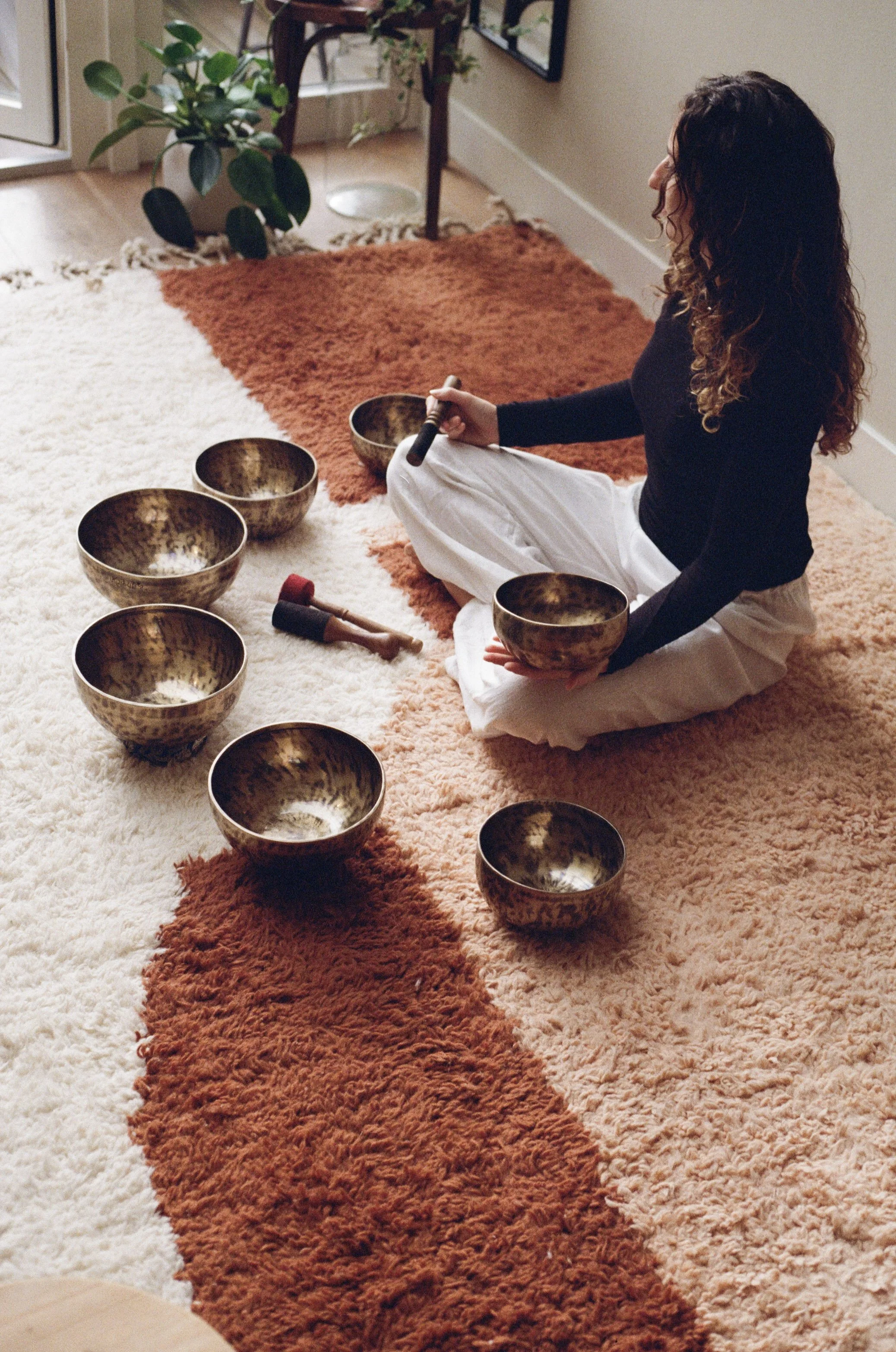 A woman sitting on a multi-colored rug, holding a tibetan singing bowl and a mallet, surrounded by several other crystal singing bowls. She is facilitating a team building workshop