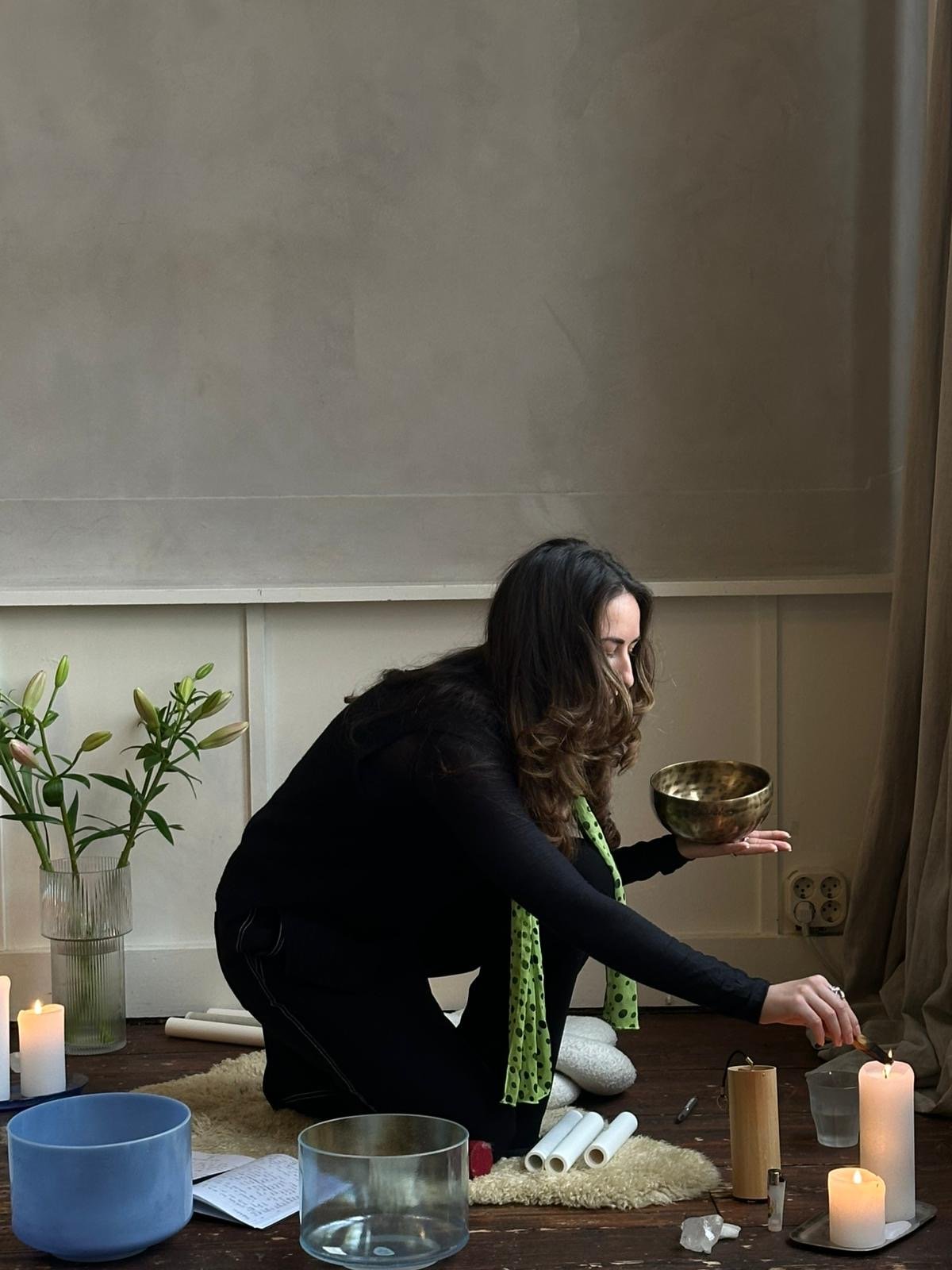 A woman with long brown hair kneeling on a cushion on the floor, holding a crystal singing bowl in her left hand and a mallet in her right hand, with lit candles and various objects around her, practicing pranayama breathing techniques & Wim Hof.