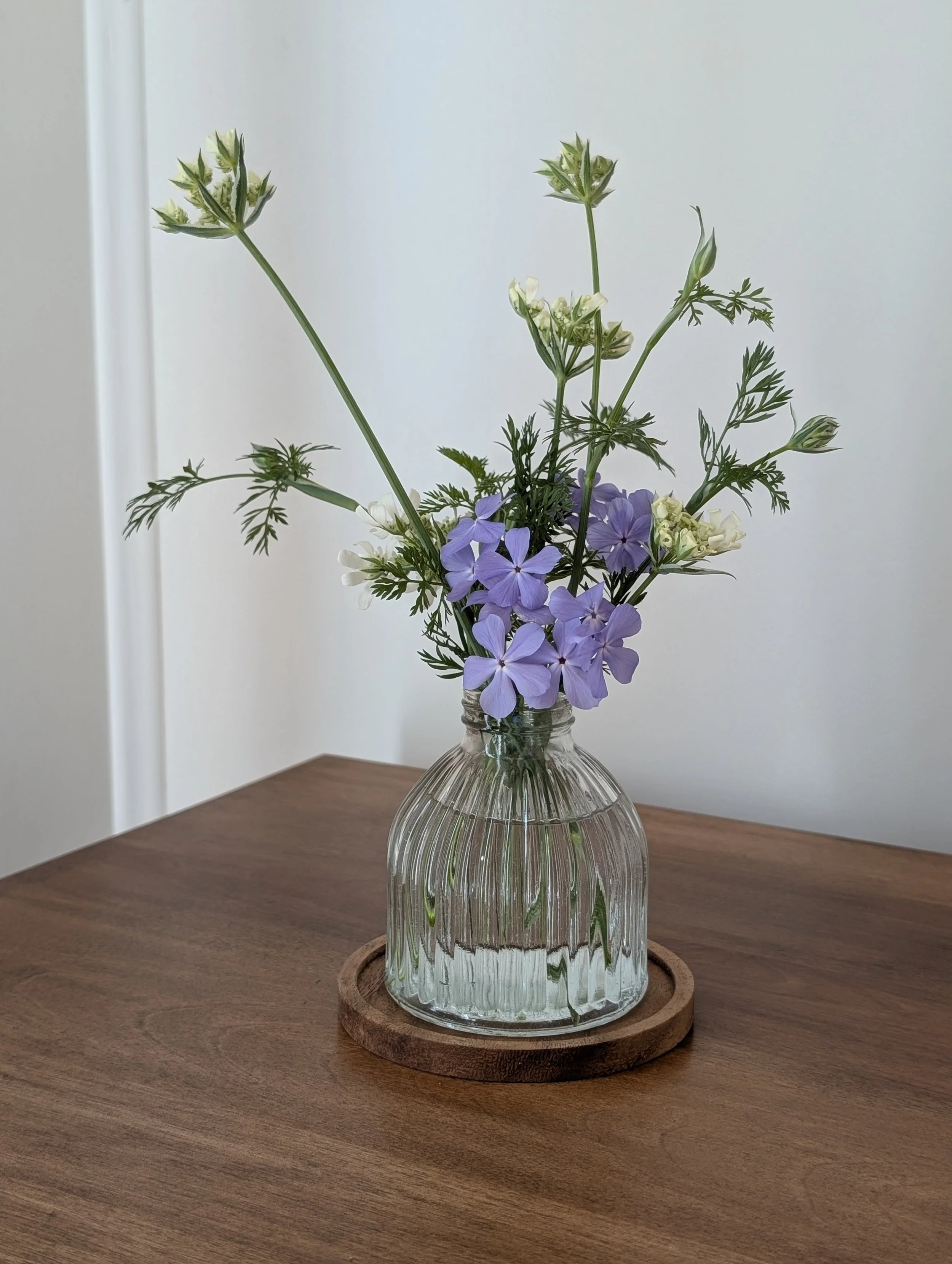 Small glass vase filled with purple and white garden flowers arranged on a wooden table in natural window light.