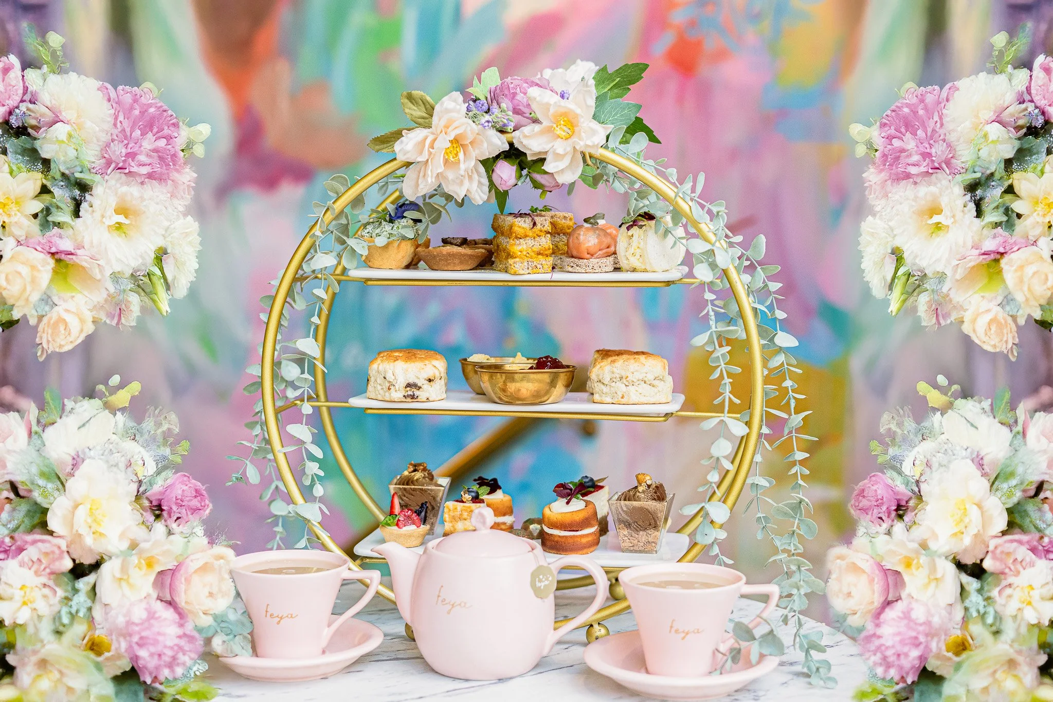 A tiered dessert display with various pastries, cakes, and sweets, surrounded by pink and white flowers and greenery, with a pastel colorful background.