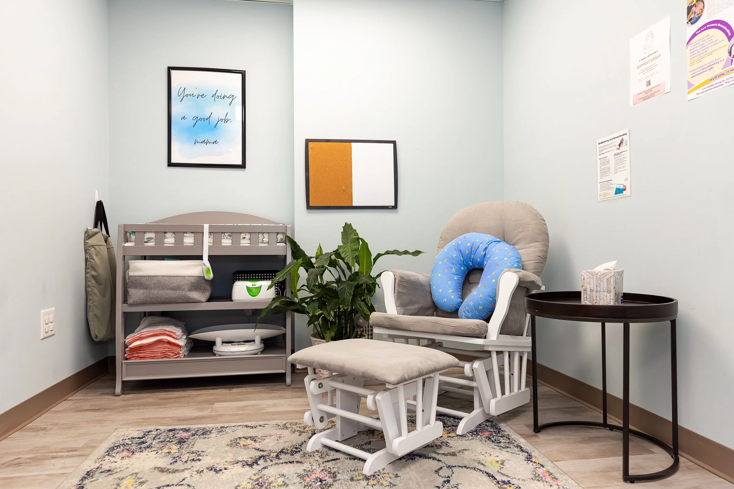 It has a beige rocking chair with a gray cushion and a blue pillow, a small beige cushioned footstool, a black round side table with a tissue box, a gray shelf with various medical supplies, a green plant, framed wall art including a motivational message, and informational posters on the light blue walls.