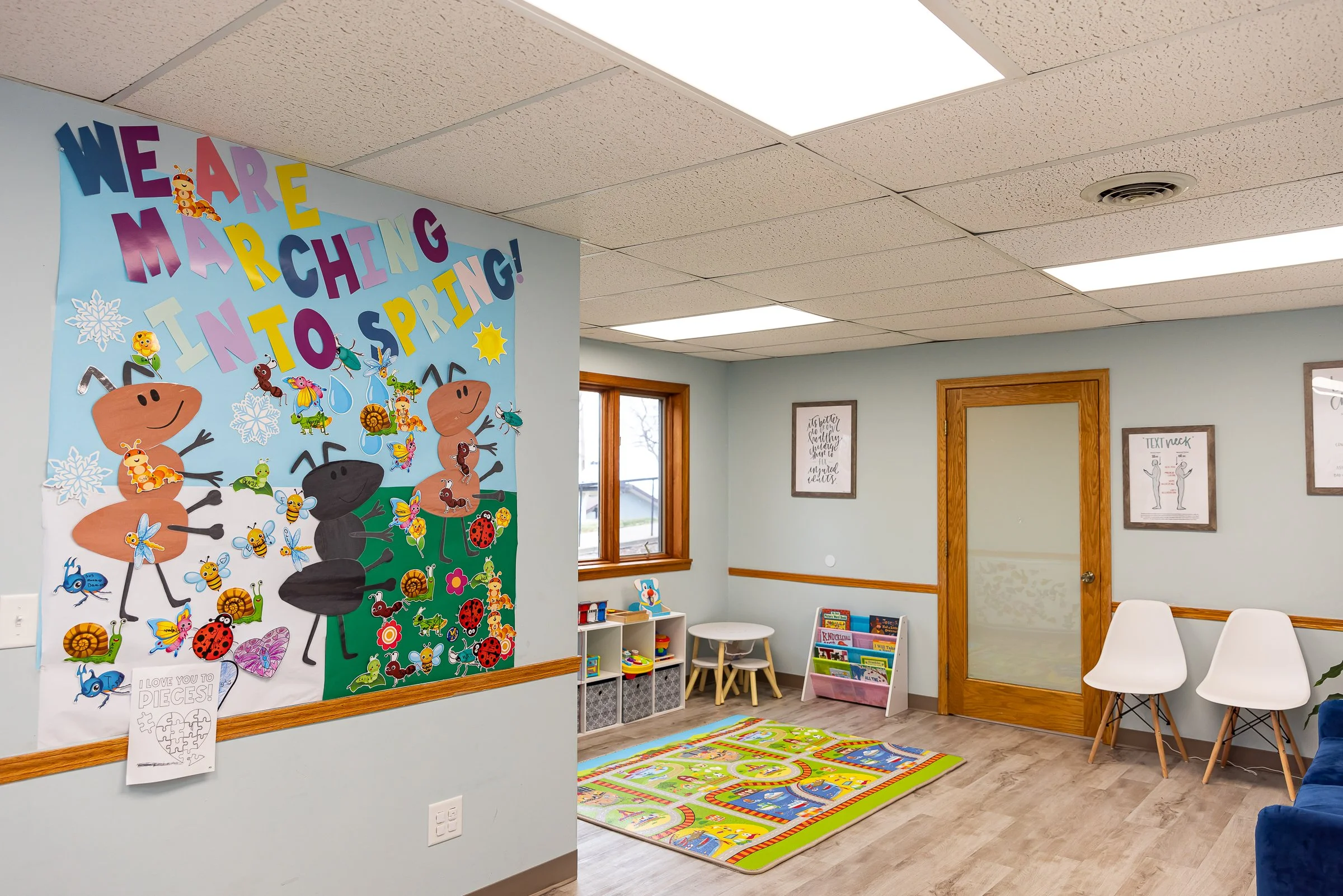 A room in a children's waiting area with a colorful spring-themed mural on the wall, a window, a small bookshelf with children's books, a play rug, and seating for children and parents.