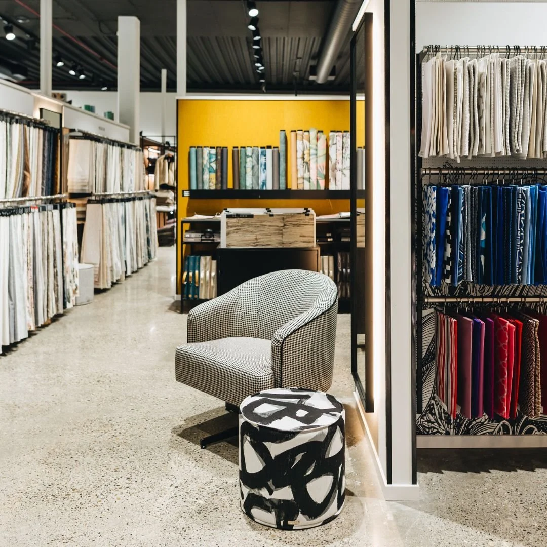 Interior of a fabric or home decor store with fabric rolls on shelves and a patterned armchair and small table near the aisle.