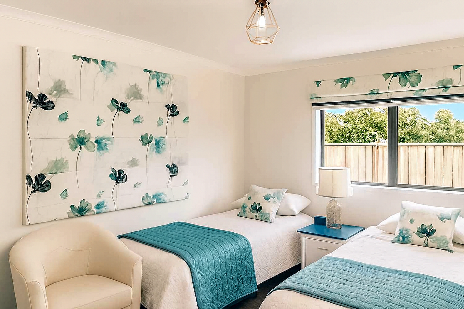 A bedroom with two twin beds, each with white bedding, teal bed scarves, and floral pillows. A nightstand with a lamp is between the beds. A large window shows a wooden fence and trees outside. There is an armchair in the corner and a wall art piece with a floral design behind the beds.