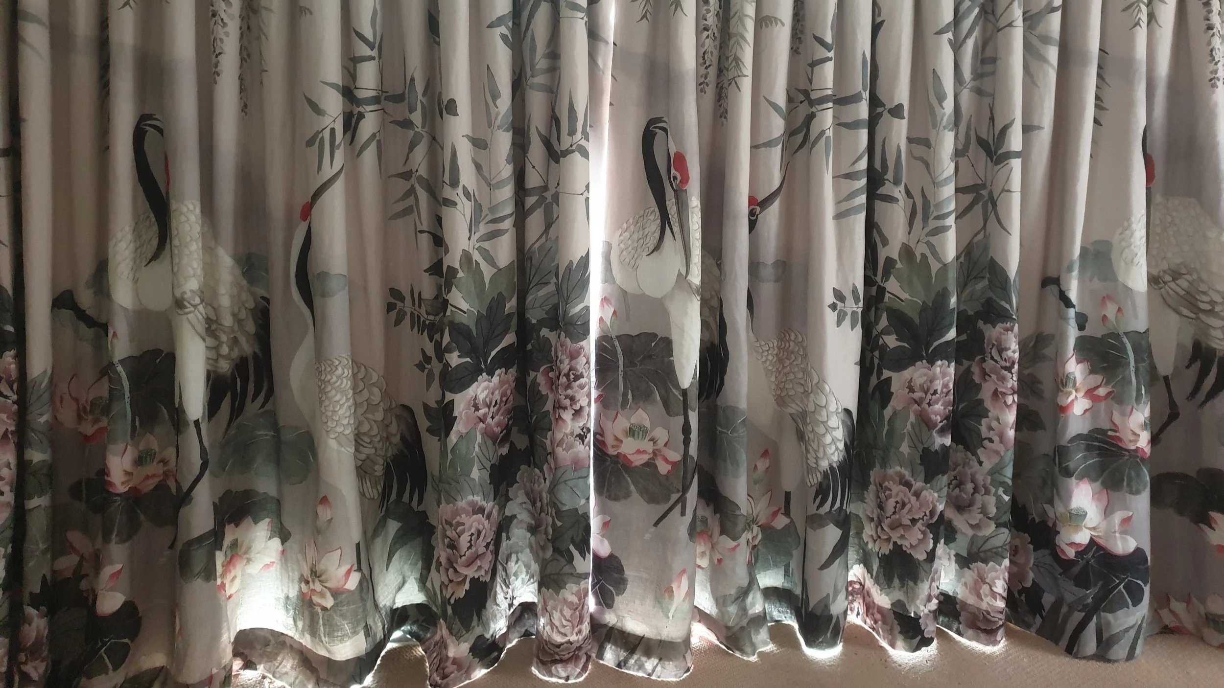 Curtains with a floral and crane bird pattern hanging in front of a window, with sunlight filtering through.