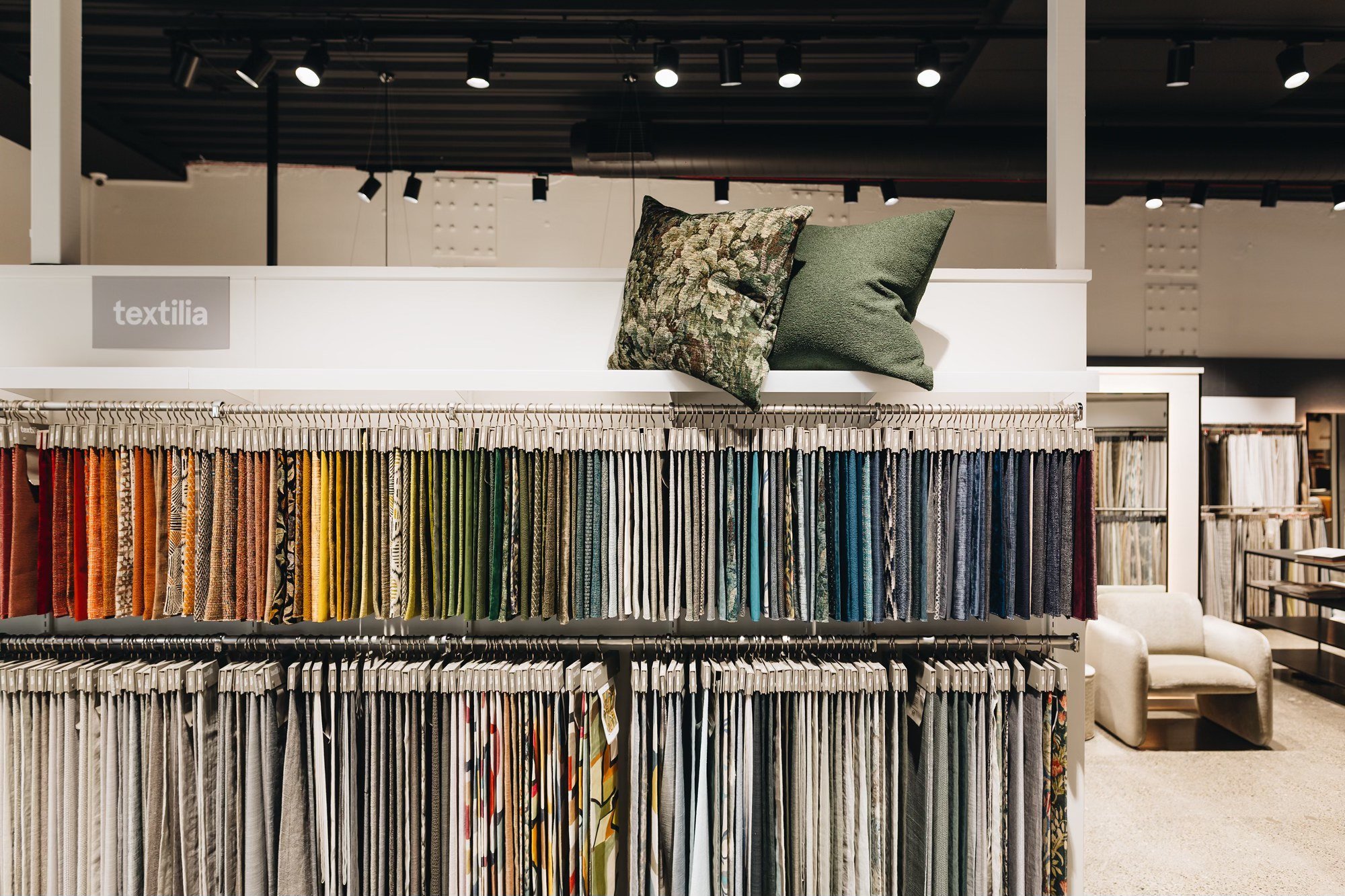 Display of fabric swatches in various colors with two decorative pillows on top, in a store with seating area in the background.