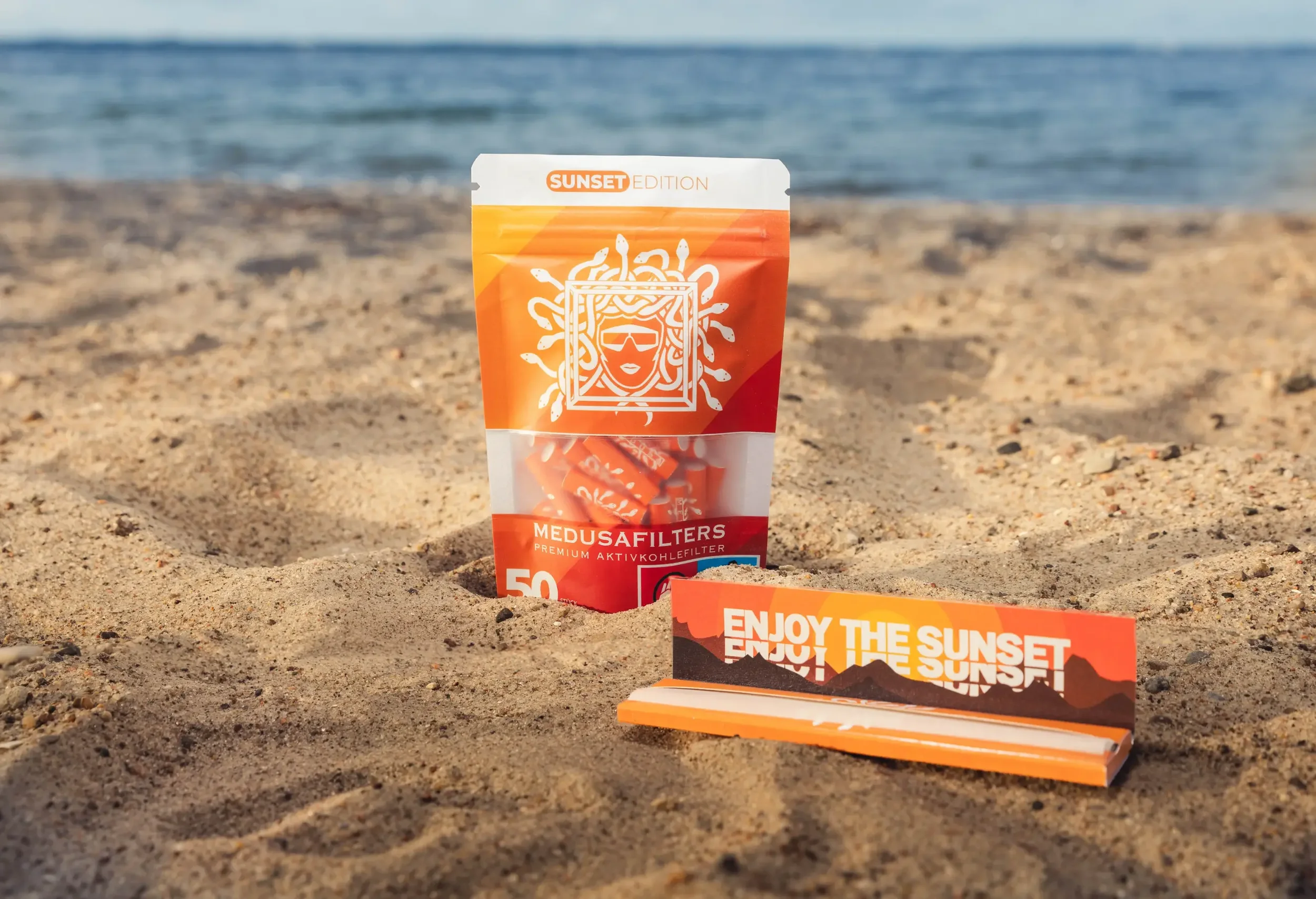 Ein Beutel mit Sonnencreme am Sandstrand mit Meer im Hintergrund. Der Beutel trägt die Aufschrift "SUNSET EDITION" und zeigt ein grafisches Design einer Sonnenbrille und einem Gesicht. Es sind auch Werbung und Informationsmaterialien auf dem Sand sic