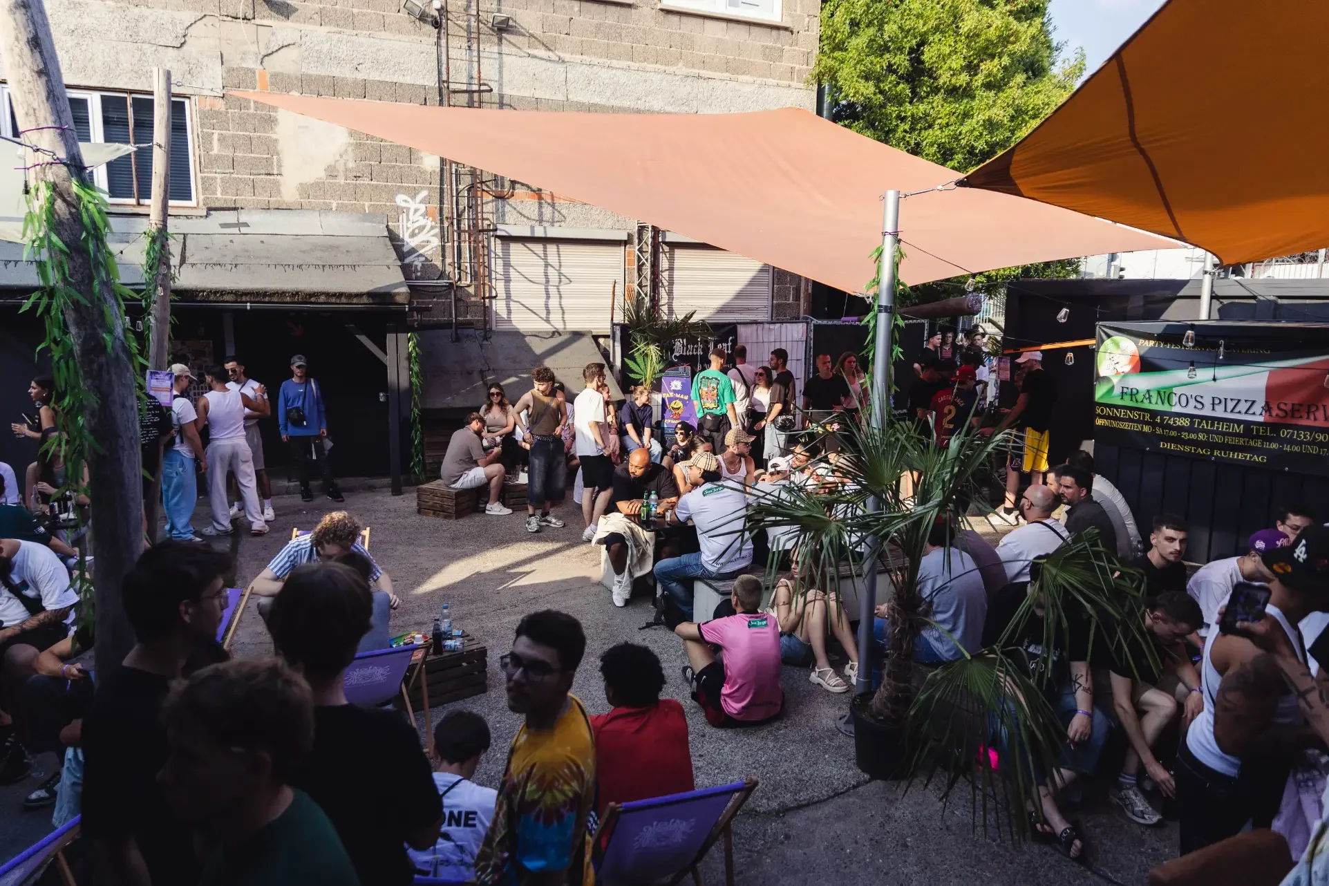 Menschen sitzen und stehen in einem belebten Außenbereich, vermutlich auf einer Veranstaltung oder in einem Café, mit Sonnensegeln und Pflanzen, im Hintergrund eine Wand mit Fenster und Graffiti.