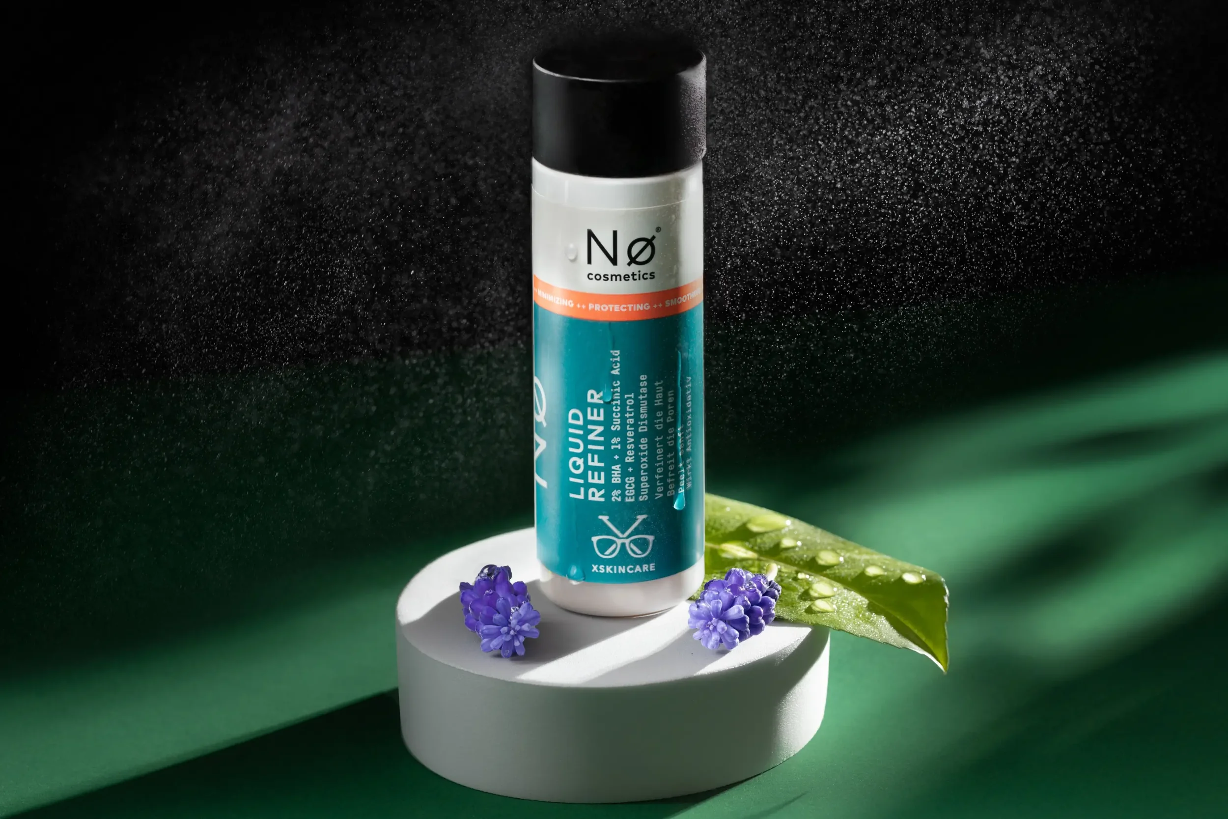 Ein Pflegeprodukt in einem blauen und weißen Behälter steht auf einem weißen Podest, umgeben von Lavendelblumen und einem grünen Blatt. Der Hintergrund ist schwarz mit Wassertröpfchen, das in die Luft sprüht.