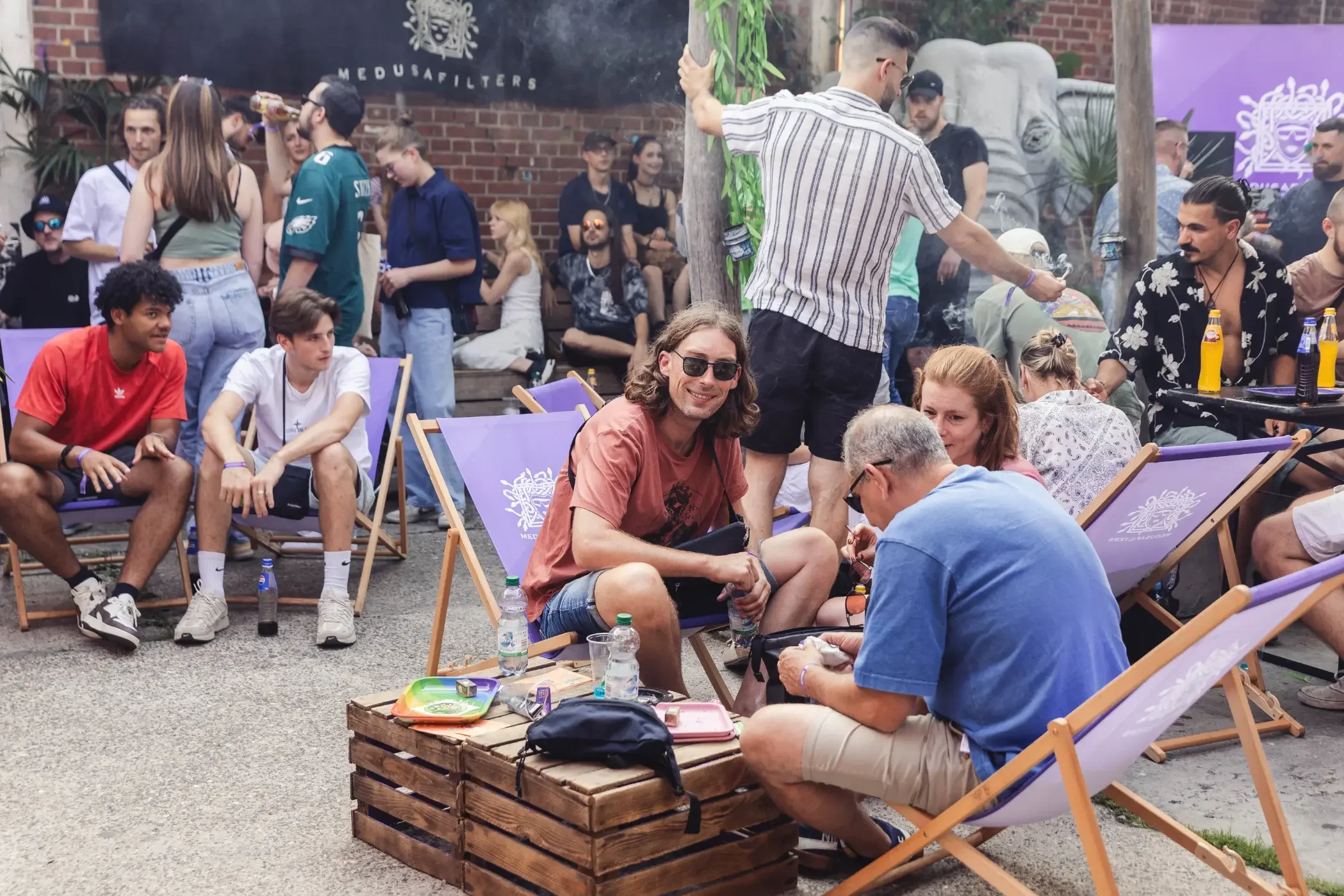 Menschen sitzen und stehen im Freien bei einem gesellschaftlichen Treffen, einige sitzen auf Klappstühlen, während andere stehen. Es gibt eine Bar mit Getränken und eine hölzerne Paletten-Tischplatte mit Trinkflaschen. Im Hintergrund ist eine Gruppe 