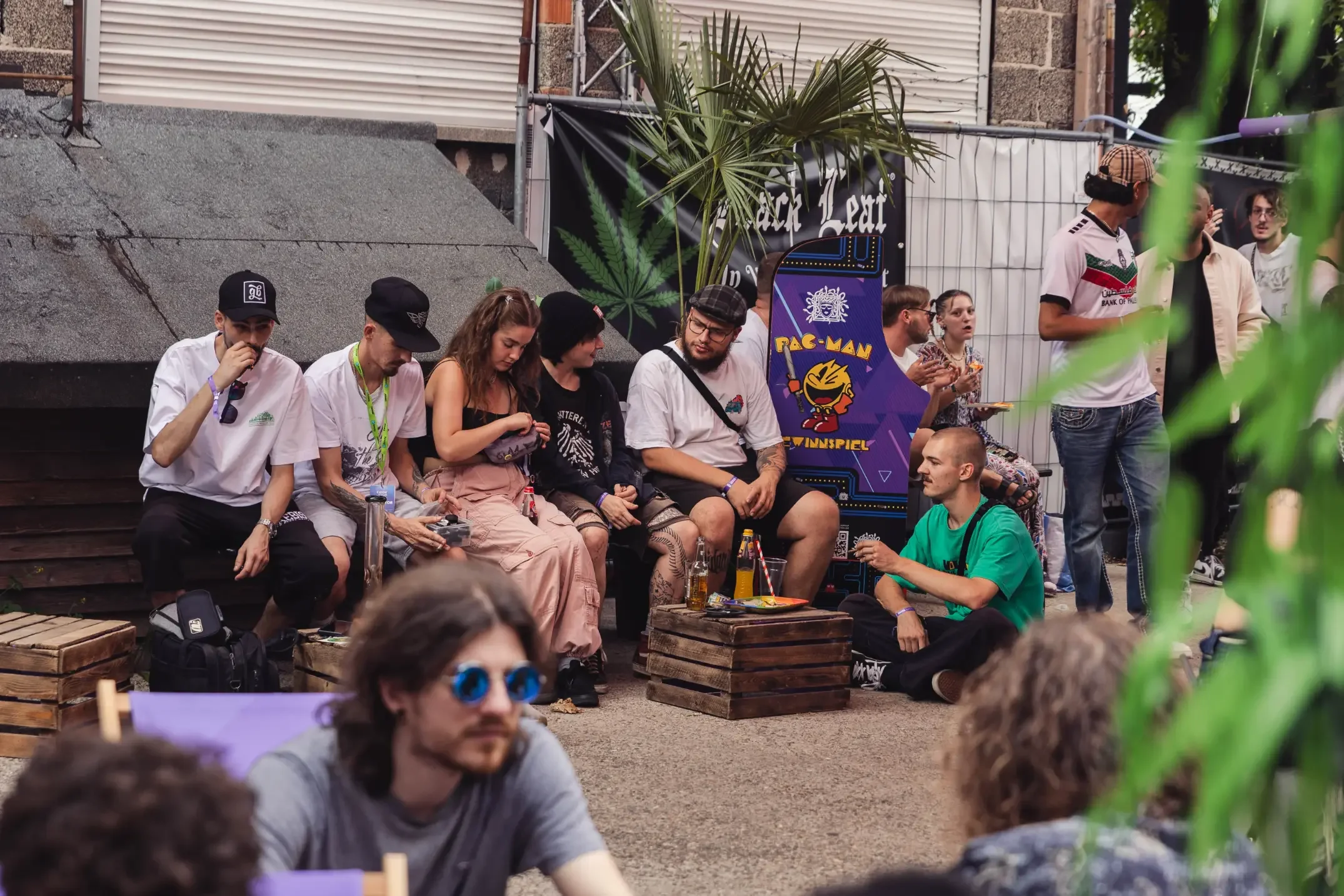Menschen sitzen im Freien bei einer Veranstaltung, es gibt eine Street-Food-Statue und einen Pac-Man-Spielautomaten, einige Leute haben Getränke in der Hand, im Hintergrund eine Palme und ein schwarzes Banner mit einem Marihuana-Blatt.