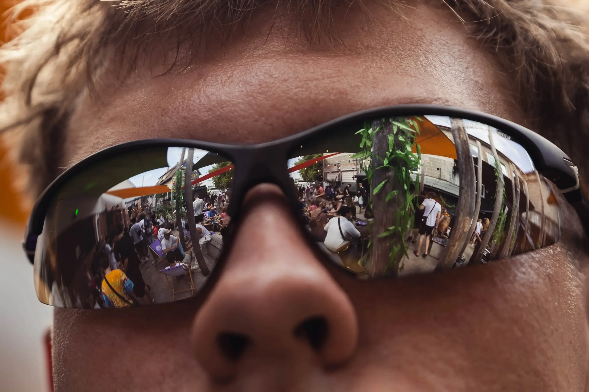 Nahaufnahme einer Person mit Sonnenbrille, die die Reflexion eines belebten Marktplatzes oder Festivals zeigt.